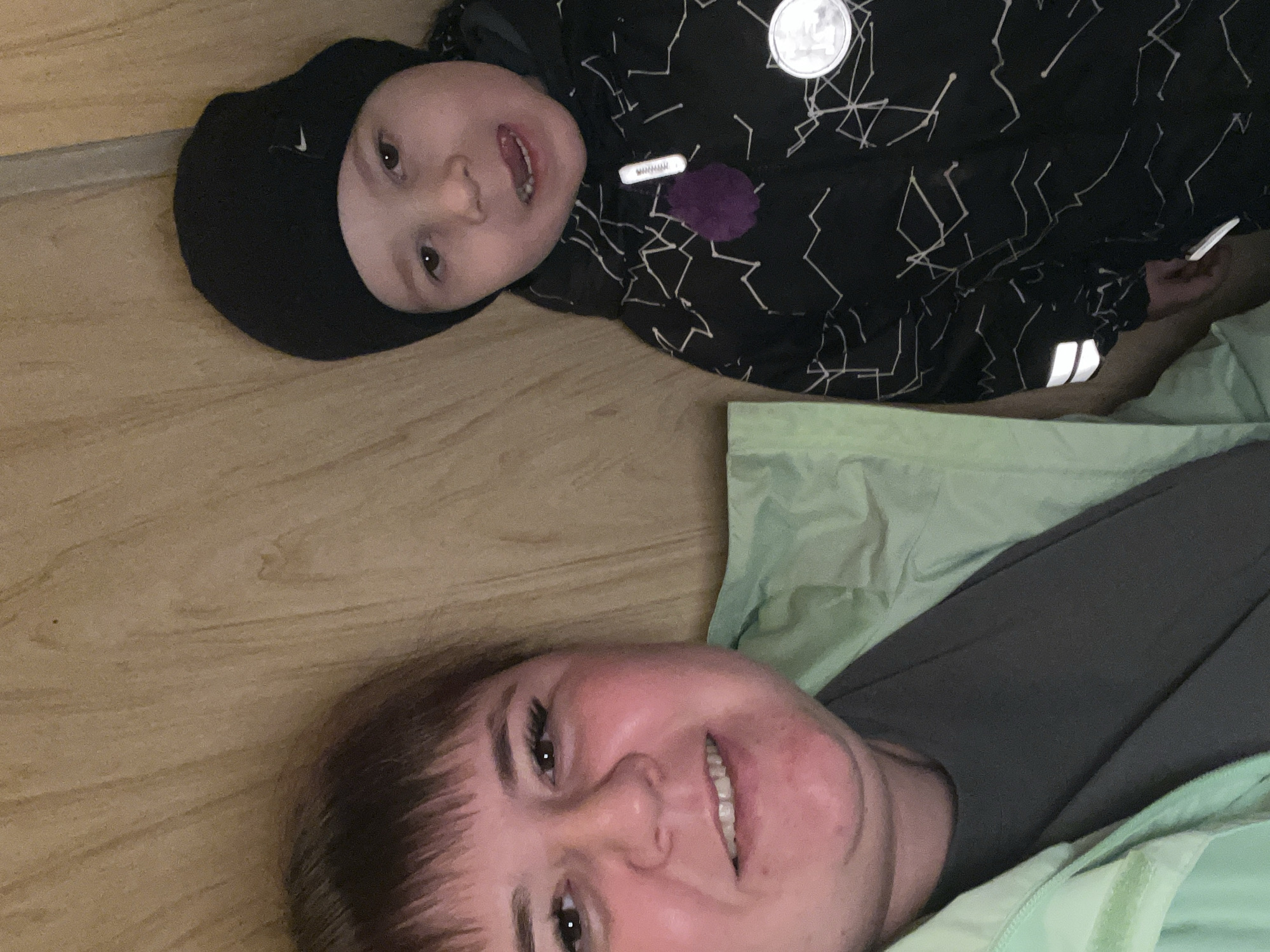 A woman and a young child are lying on a light-colored wooden floor. The woman is in the lower portion of the frame, smiling with her face angled towards the camera. She is wearing a dark gray shirt and a light green jacket. The child is positioned above her, also looking towards the camera and smiling. The child is wearing a black beanie with a white logo, and a black jacket with white zigzag patterns. The jacket has a round metallic emblem, a white rectangular tag, a purple pom-pom, and a reflective stripe on the sleeve. The location is indoors, possibly a home or apartment in the city of Chaikovsky, Russia.