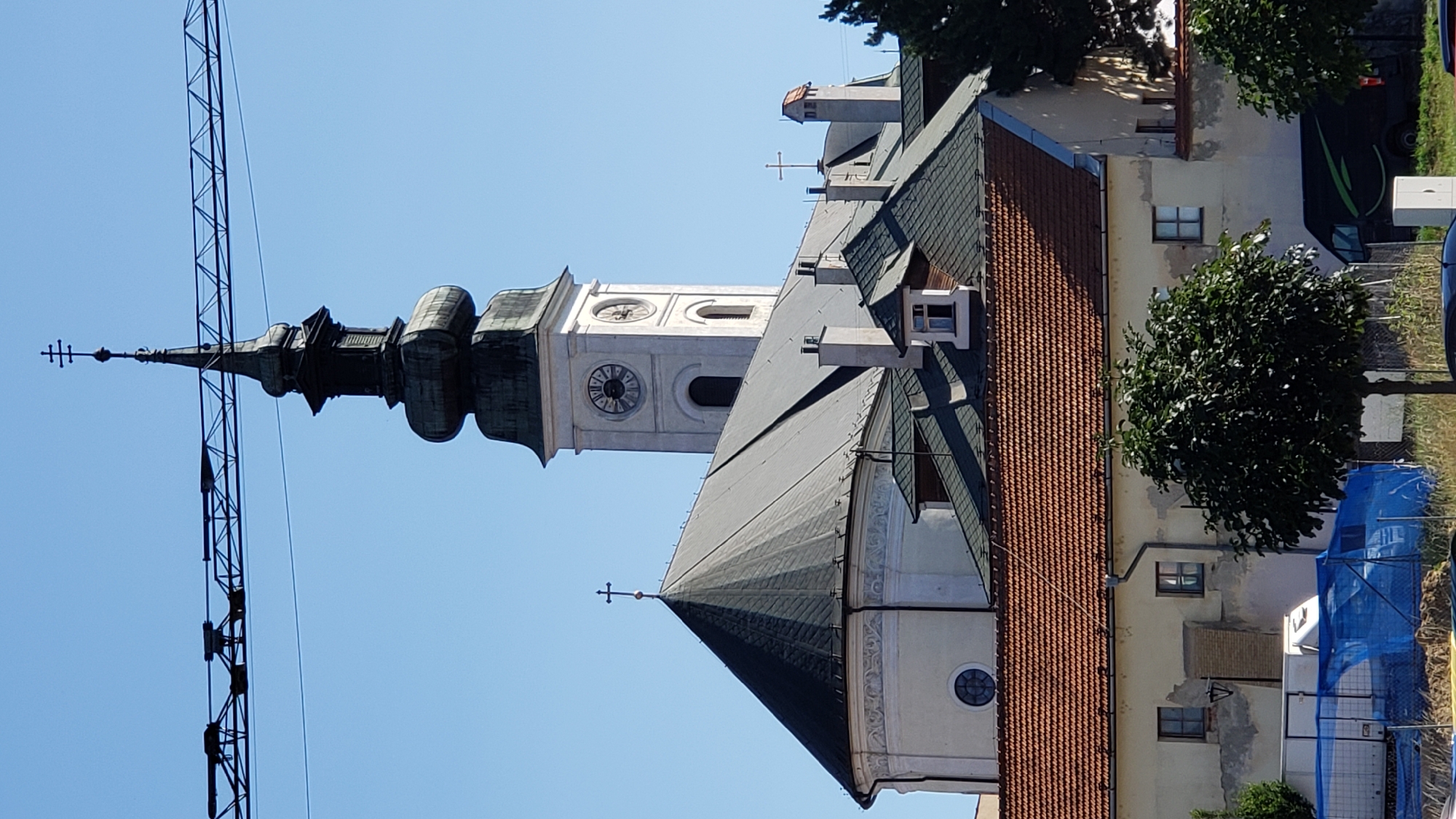 The image shows a view of the St. Nicholas Cathedral in Prešov, Slovakia. The cathedral's prominent clock tower, topped with a distinctive spire and cross, is visible against a clear blue sky. The main building of the cathedral features a large, dark-tiled conical roof. In the foreground, several buildings with red-tiled roofs are partially visible, along with trees and a construction crane extending across the upper portion of the frame. A blue tarp covers some construction materials in the lower left. The scene appears to be a bright, sunny day.