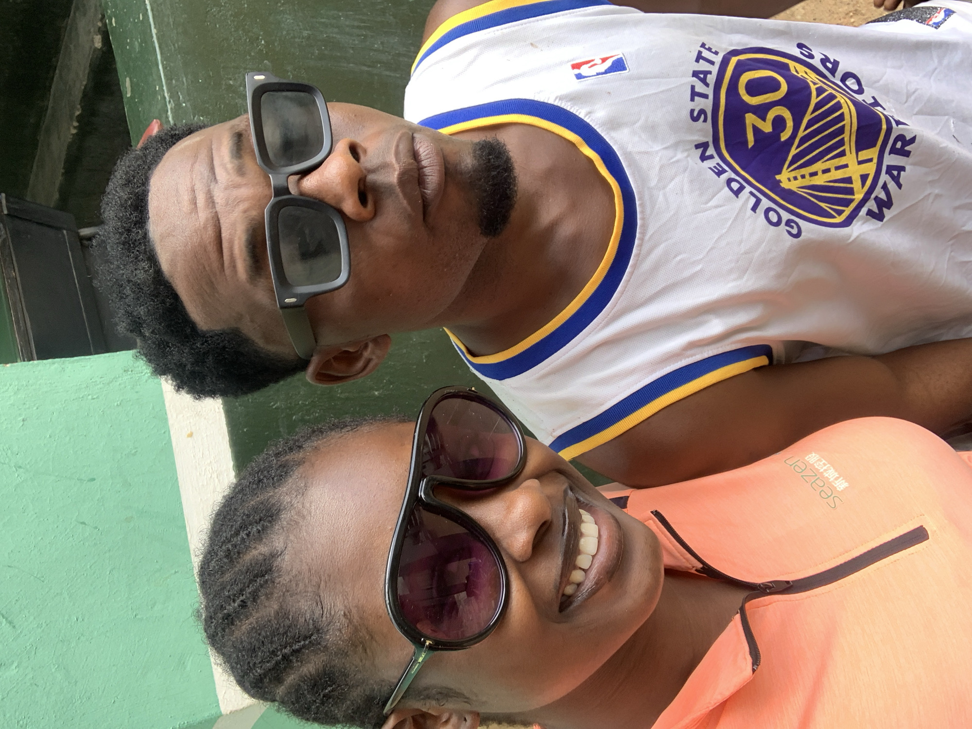 Two individuals are closely positioned; one wears a white jersey with yellow and blue trim, featuring a Golden State Warriors logo and the number 30. The other person wears an orange top with a visible zipper and logo, and dark sunglasses. Both individuals are dark-skinned, and the background suggests an outdoor environment. Location is in the city of New Stadium, Nigeria.