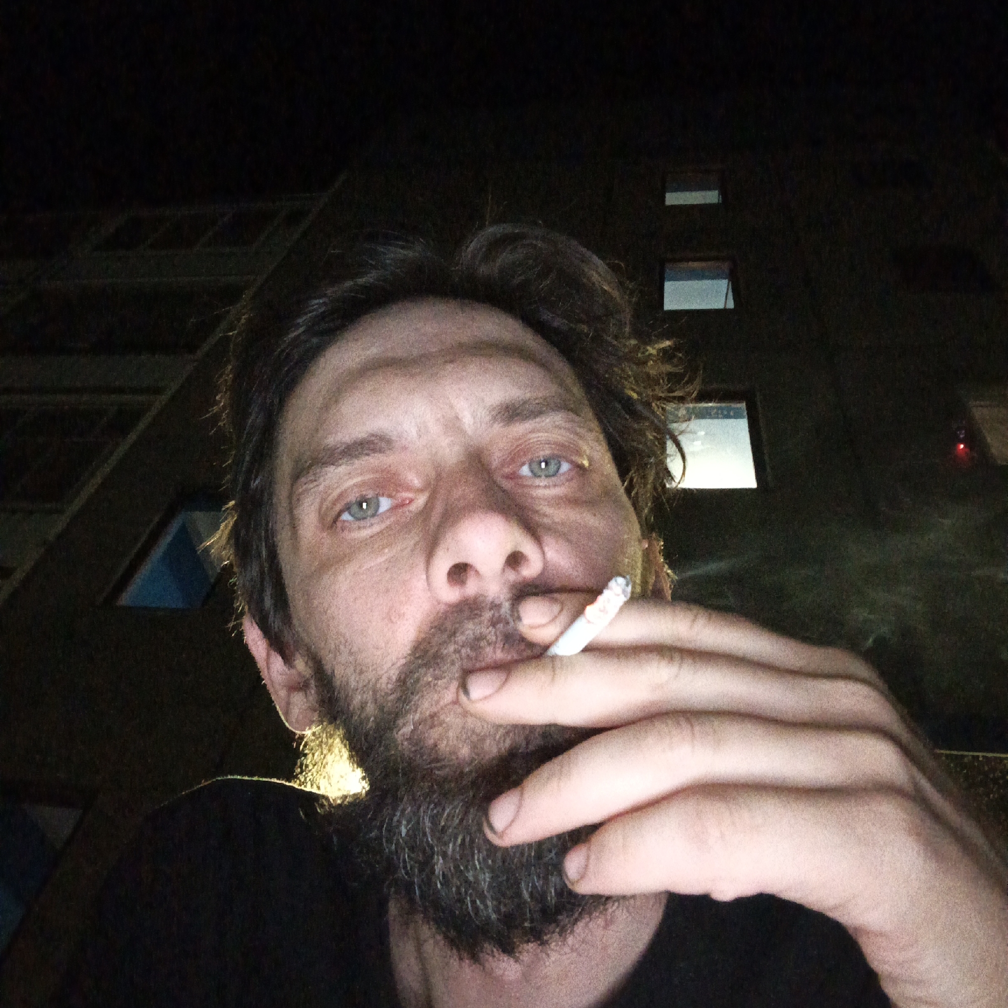 A low-angle, close-up portrait of a Caucasian man with a beard, holding a cigarette in his mouth and cupped in his right hand. The man has messy dark hair, and light-colored eyes. He is wearing a black shirt. The background shows the facade of a tall building, with visible windows and balconies, in a dark environment suggesting it is night.
It is not possible to determine the location as the city of Марковский, Russia using only the image, as that is not directly discernible from the visual information provided.