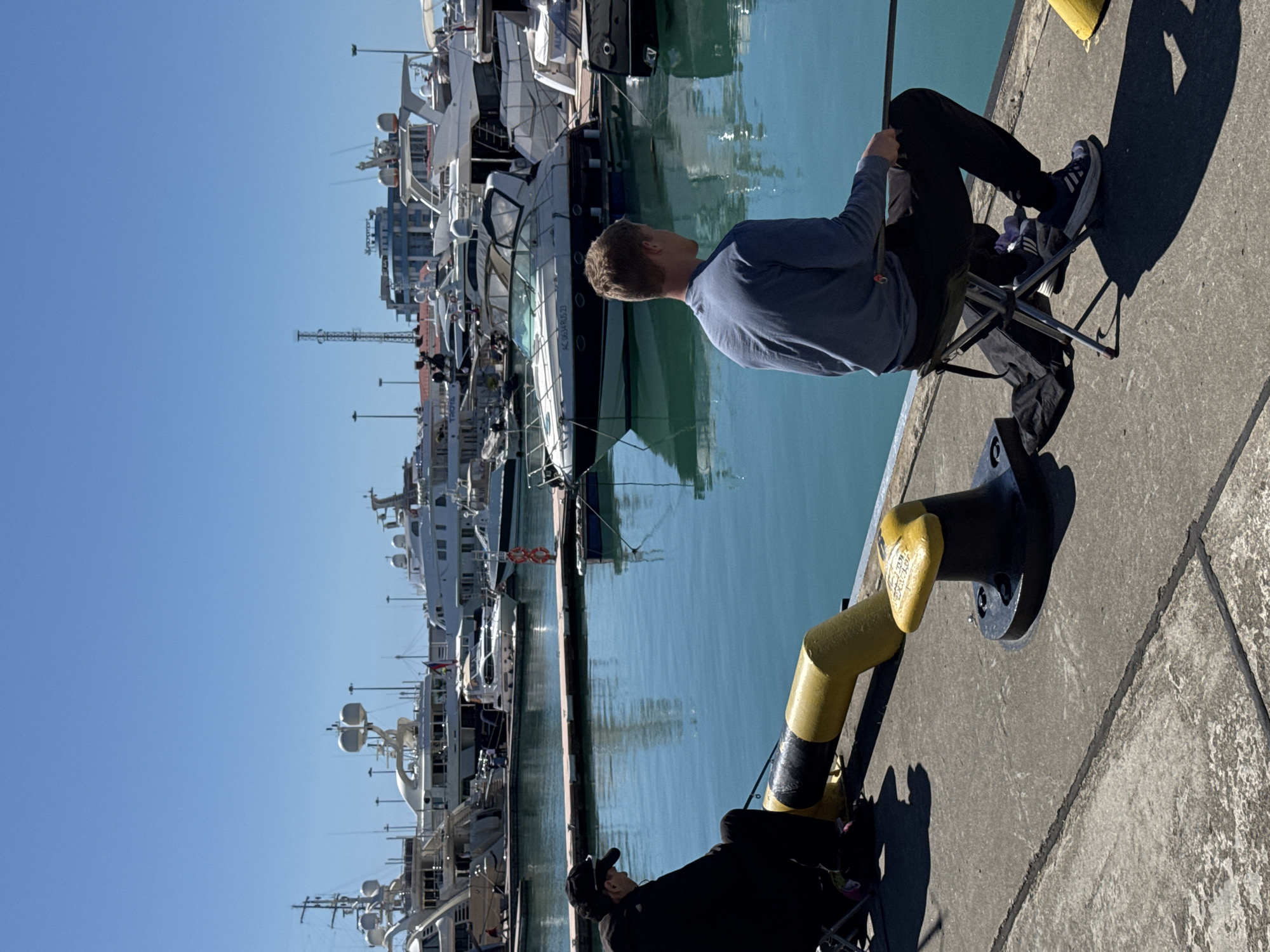 The image captures a sunny day at a marina, likely in Sochi, Russia, with two men relaxing by the water amidst numerous docked yachts. The scene is dominated by a clear blue sky and calm, turquoise-green water.
In the foreground, two men are seated on folding chairs on a concrete dock. The more prominent man, positioned closer to the viewer, wears a blue long-sleeved shirt, dark pants, and athletic shoes. He is seen in profile, looking towards the array of boats. To his left, partially visible, another man in a dark jacket and cap is also seated, seemingly observing the surroundings. Both men are casting long shadows on the concrete, suggesting a mid-morning or late afternoon sun.
Behind the men, a long line of white and dark-hulled yachts of varying sizes is docked. The boats are reflected clearly in the still water. Many of them feature multiple decks, antennas, and radar domes. One particular yacht has text visible on its hull: "HC 0834-0010 23". Further in the background, some urban structures and what appears to be a crane or tower are visible above the yachts. Yellow and black striped bollards and mooring fixtures are present along the edge of the dock, one of which appears to have faint, unreadable text. The overall atmosphere appears tranquil and relaxed.