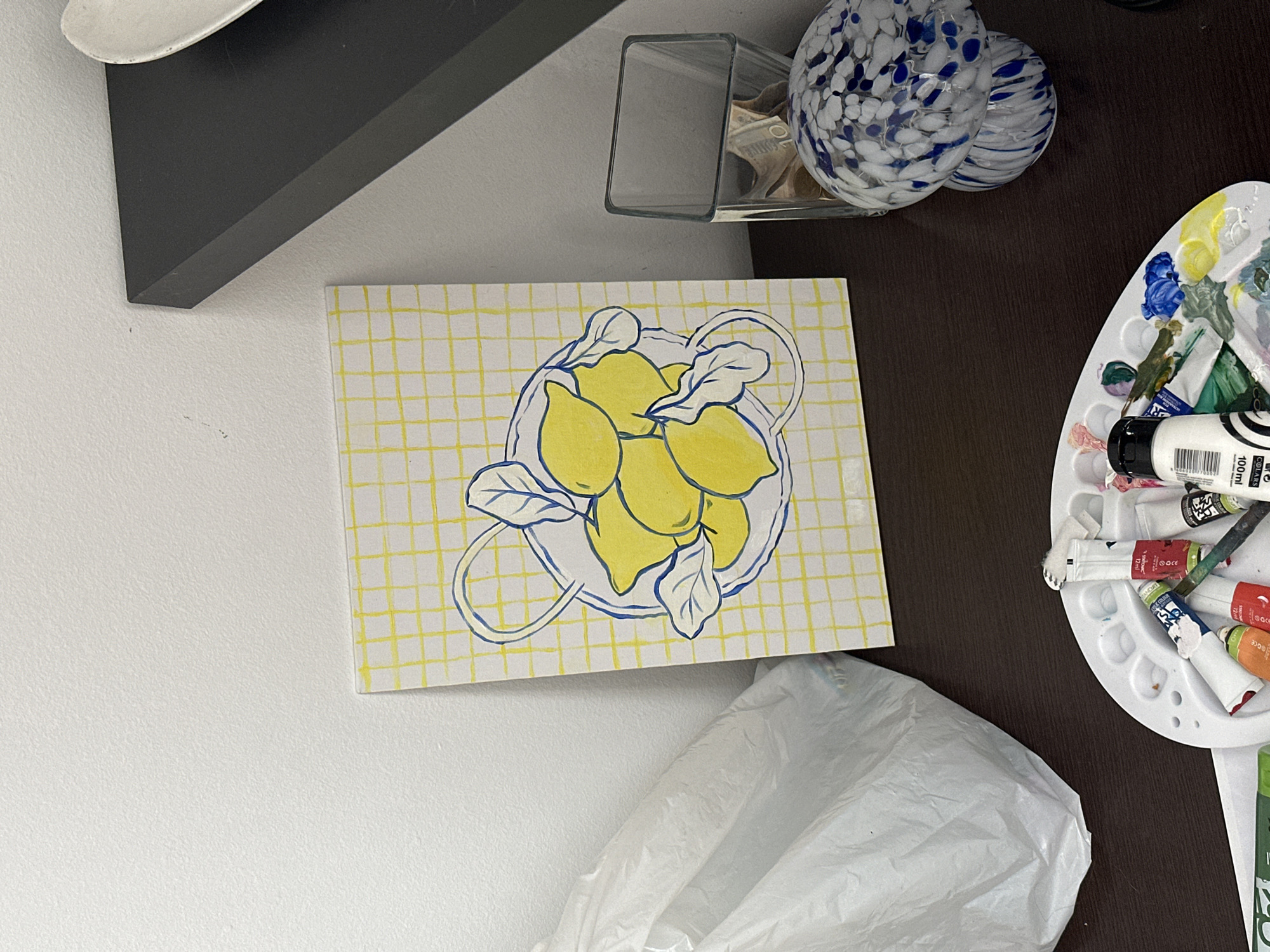 The image shows a tabletop scene with various objects. A framed painting depicting lemons on a yellow grid sits on a white surface adjacent to a dark brown tabletop. Above the painting are a glass container with unidentified contents and two blue and white patterned orbs. Adjacent to the painting on the brown tabletop is a white painter's palette with various paint tubes and used paint. A crumpled white plastic bag is visible beneath the painting and palette. The environment consists of a white wall. There is no visible location in Novi Sad, Serbia.