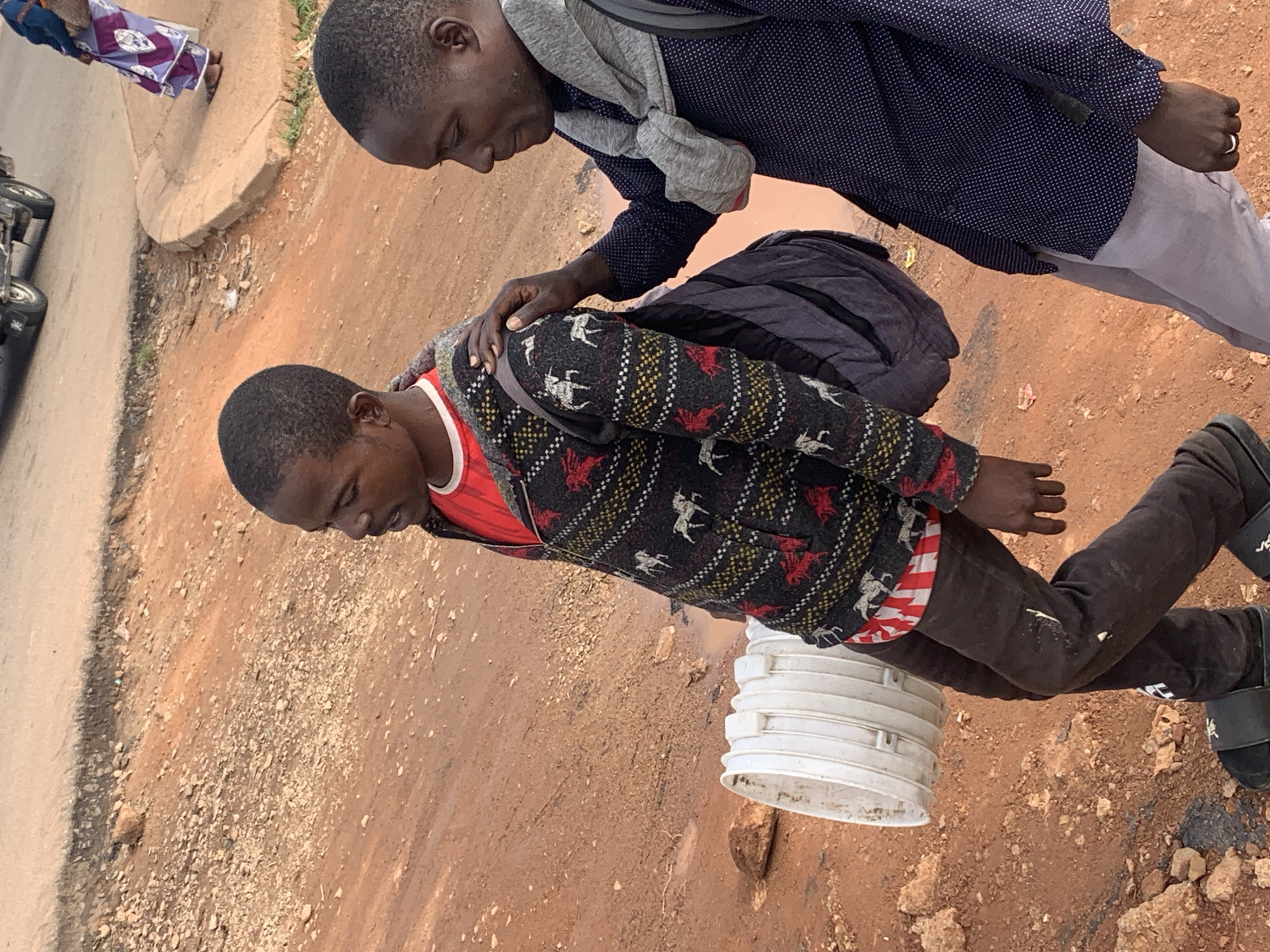 Two Black men are walking along a dirt road in Gura Top, Nigeria. The man on the left is wearing a red shirt, a patterned sweater, and dark pants. He is carrying a large white bucket. The man on the right is wearing a dark blue polka dot shirt and light gray pants. He has his hand on the shoulder of the man on the left. In the background, there is a paved road with a vehicle, and a woman in a colorful dress is walking on the sidewalk. The environment is rural, with reddish-brown dirt and scattered rocks.