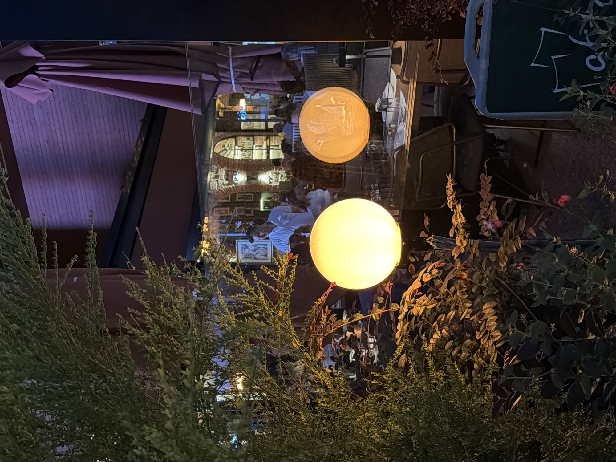 This image captures an evening scene through a glass pane, likely a restaurant or cafe window, in Kadıköy, Turkey. In the foreground, lush green foliage with some reddish-brown leaves partially obscures the view. Two spherical lamps are prominent: one is a bright, yellow-white orb on the left, and the other, to its right, is an orange orb with a carved design depicting a sailboat.
Behind the foliage and lamps, the interior of the establishment is visible. People are seated at tables, some appearing to be dining or socializing. The architecture suggests a restaurant with a warm, inviting ambiance, featuring exposed brickwork and arched doorways. A man in a white shirt is standing and looking towards the camera, or the viewer. A large, folded pink umbrella hangs from an overhead structure on the right side of the frame, suggesting an outdoor seating area or a covered walkway. The overall lighting is dim and atmospheric, with the lamps providing focal points of light. The scene is likely set on a street or a pedestrian area in Kadıköy, a vibrant district known for its cafes and social spaces.