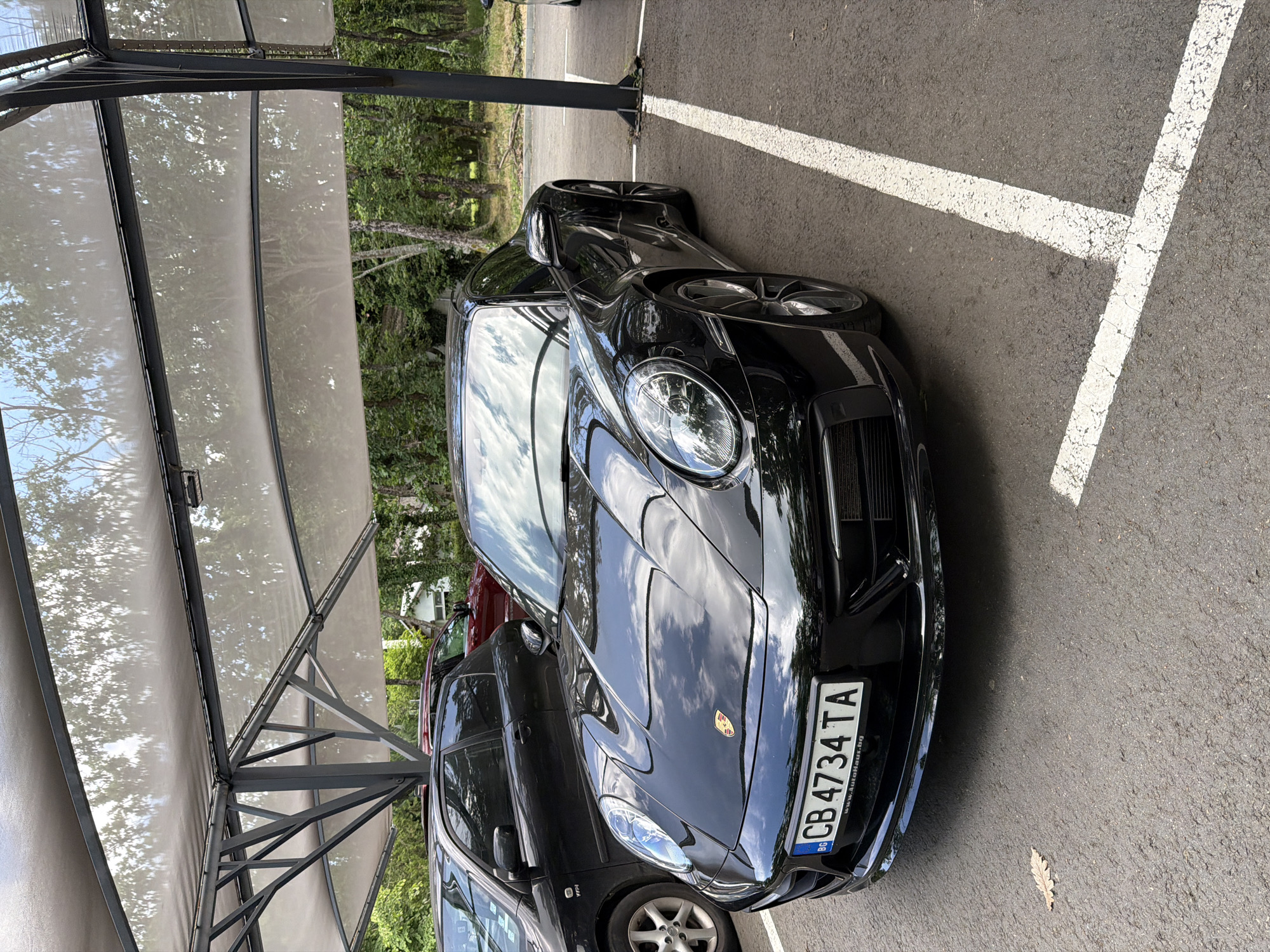 A black Porsche sports car with the license plate CB 4734 TA is parked in a parking spot with white lines on asphalt. The car is partially under a covered structure with a fabric roof supported by metal beams. The environment appears to be an outdoor parking area, with trees visible in the background. Another car is parked alongside the Porsche. The license plate suggests the car is registered in Bulgaria. There is no evidence to suggest a location in Burgas, Bulgaria.