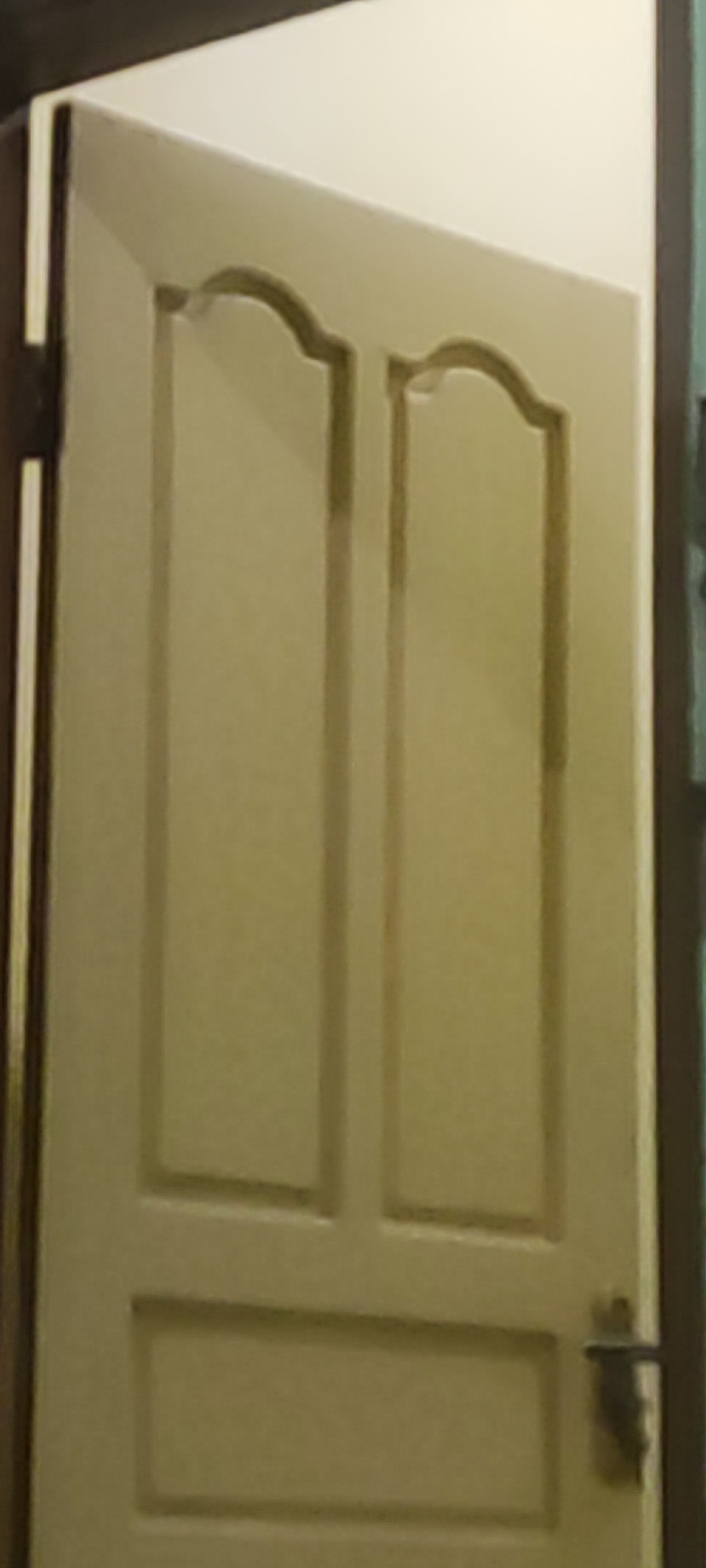This is a close-up, slightly blurred image of a beige interior door. The door has two raised panels on the upper portion, each with an arched top. Below the panels, there is a horizontal raised rectangle. A dark-colored door handle is visible on the right side of the door. The door appears to be slightly ajar. The image does not provide any contextual clues to determine its location in the city of Gebang, Indonesia.