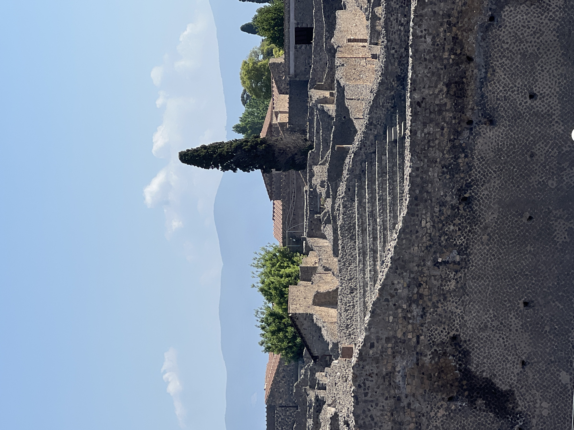 The image shows a panoramic view of the archaeological site of Pompeii, Italy, under a clear blue sky with scattered clouds. The foreground features the weathered stone structures of ancient Roman buildings, characterized by rough-hewn stone walls and remnants of staircases leading up through the ruins. These structures exhibit a layered appearance, with different levels of construction visible. In the mid-ground, more well-preserved buildings with terracotta-tiled roofs are interspersed with lush green trees, including a tall, dark evergreen tree that stands out prominently. Further in the background, hazy blue mountains form a natural horizon, with a few wispy clouds dotting the sky. The overall scene depicts the historical ruins of Pompeii, showcasing its ancient architecture and the surrounding natural landscape.