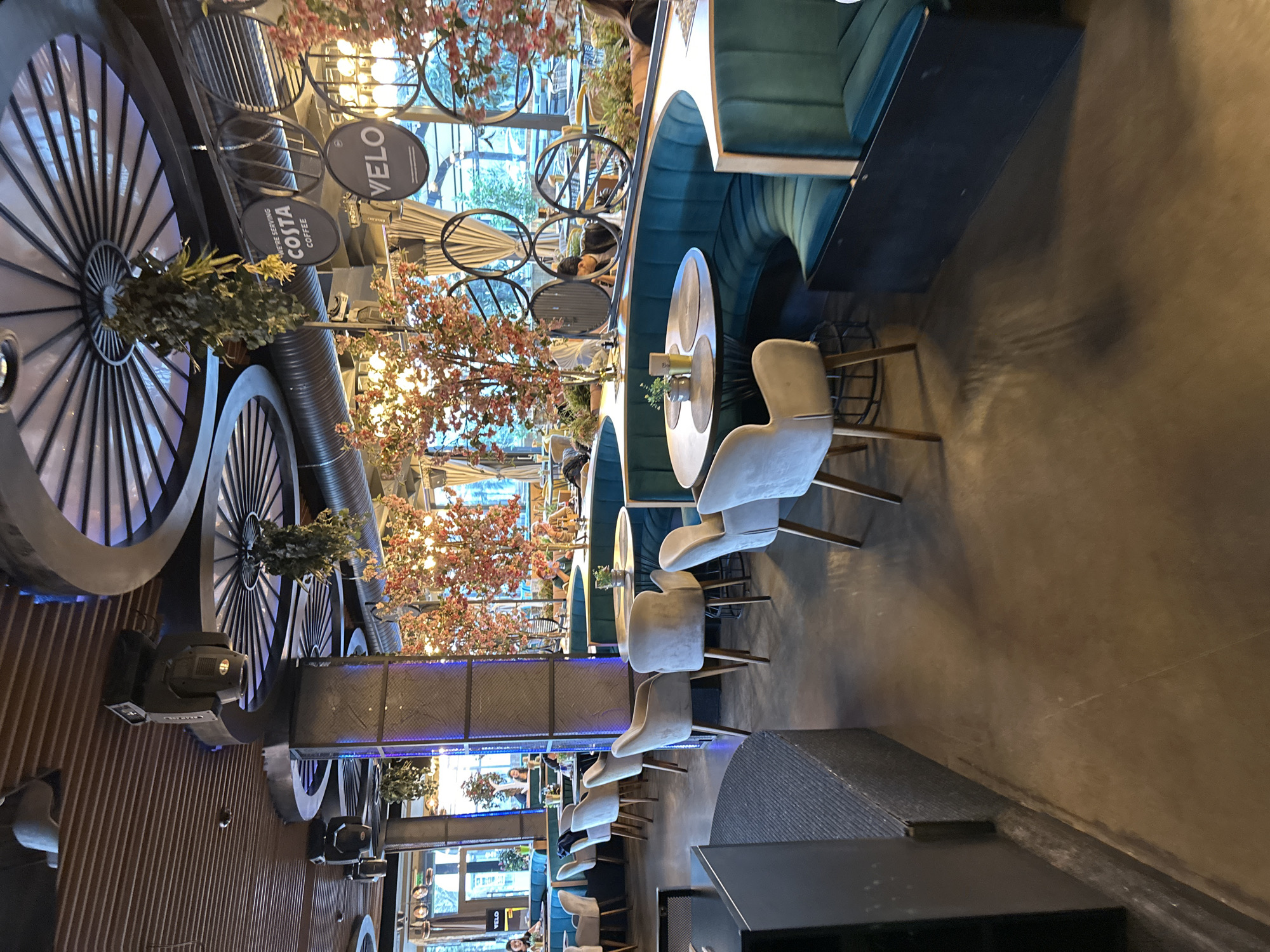 The image shows the interior of a café in Sofia, Bulgaria. The interior design includes decorative elements of large, stylized wheels on the wall and smaller bicycle wheels suspended from the ceiling. There are seating areas with curved, green upholstered booths and round tables, each with a set of chairs. People are seated at some of the tables. The café has large windows that allow natural light to enter. There are also artificial lighting fixtures, including spotlights. There is a sign for "Costa Coffee."