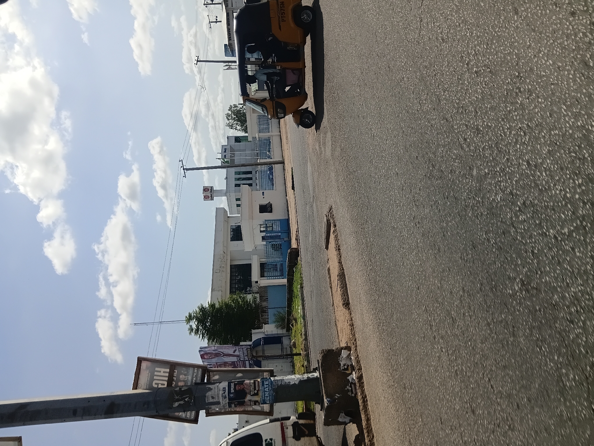 In Jalingo, Nigeria, a yellow motorized rickshaw, identified by the license plate PT575M, is moving on a paved road under a partly cloudy sky. Two individuals are seated inside the vehicle. To the left, a utility pole stands prominently, adorned with multiple posters. One poster displays "HIC" in large letters, followed by portraits. Another poster features a man in a white garment and a tie, with text that appears to include "MUHAMMAD SAUTAYA" and "GOVERNOR." Further left, a portion of a white vehicle is visible. In the background, a low-rise white building with dark windows is situated behind a blue metal fence. A small evergreen tree is planted near the building. The road surface is asphalt, with some visible wear and tear.