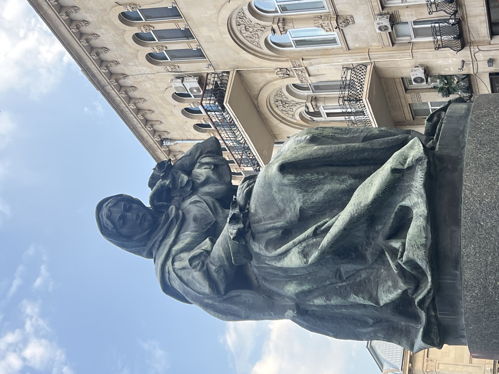A bronze statue of a woman wearing a headscarf and a long, flowing dress is positioned in the foreground, to the left of the frame. The woman is seated and appears to be holding something in her lap. Her head is tilted down, and her gaze is directed downwards. Behind the statue, a beige building with arched windows and balconies with ornate railings is visible. The building has decorative architectural details along its facade. The sky above is partly cloudy, with patches of blue visible. The scene is set outdoors in an urban environment, likely in the Sabail Raion district of Azerbaijan, given the architectural style.
