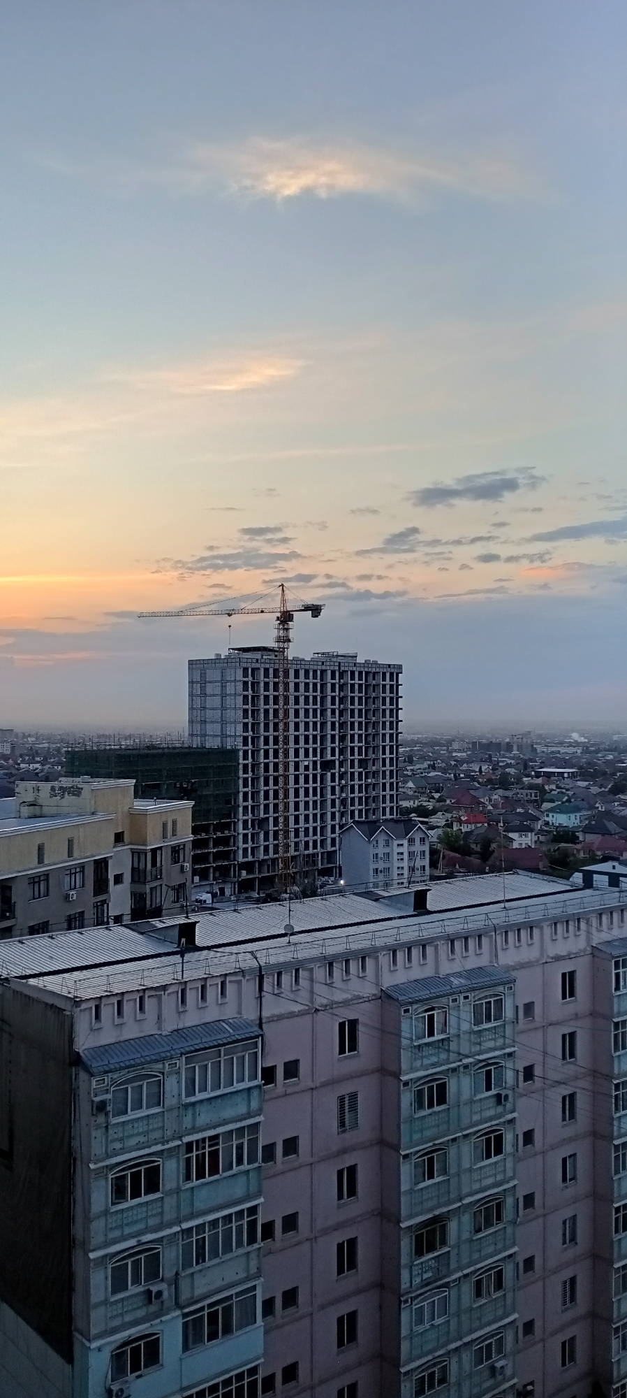 The image depicts an urban landscape at sunset in Bishkek, Kyrgyzstan. In the foreground, several apartment buildings are visible, with the closest one featuring a light blue facade and numerous balconies with glass enclosures. Behind these buildings, a large, unfinished concrete high-rise structure dominates the scene, topped by a construction crane. The sky displays a gradient of colors, from pale orange and yellow near the horizon to a soft blue overhead, with scattered clouds. The background reveals a sprawling city with more residential buildings and a hazy atmosphere, indicative of urban development and possibly dust. The overall scene captures a moment of transition from day to night in a developing city.