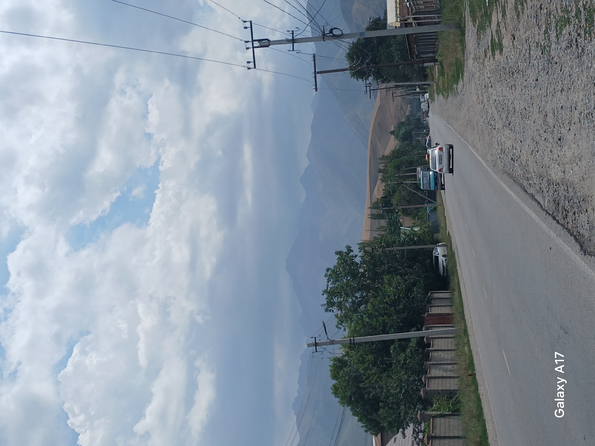 The image depicts a scene along a road in Koi-Tash, Kyrgyzstan. The sky is partly cloudy with scattered white clouds against a pale blue backdrop. In the distance, a range of muted, hazy mountains is visible. The foreground features a paved road with white lane markings. On the left side of the road, a stone wall borders a property with green trees behind it. A white car is parked on the left shoulder of the road. Further down the road, several vehicles, including a white car and a blue truck, are traveling in the same direction. On the right side of the road, utility poles with wires are prominent, and there are some structures and trees. The ground on the right is covered in gravel and grass. The overall impression is of a rural or suburban roadside environment on a moderately overcast day. The watermark "Galaxy A17" is present in the bottom left corner.