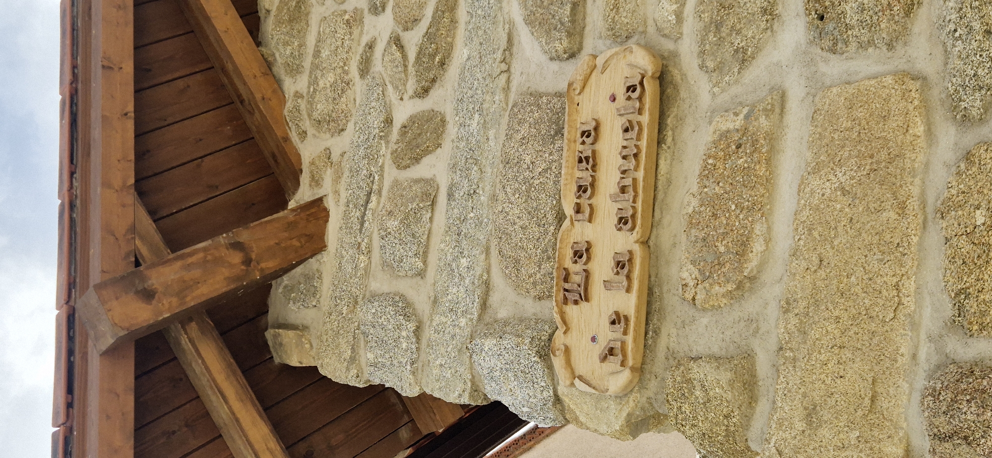 The image shows a close-up of a rustic stone wall with a wooden sign attached. The sign is carved from light-colored wood and has text engraved on it. The text appears to be in a script that resembles Cyrillic or a similar alphabet. The sign is decorated with two small red embellishments at the top and bottom. The stone wall is constructed from irregularly shaped stones of varying sizes, with mortar filling the gaps between them. The stone colors range from light gray to yellowish-brown. To the left of the stone wall, a portion of a wooden structure is visible, including beams and wooden planks, suggesting an overhang or roof. The sky is visible in the upper left corner, indicating it is daytime and possibly cloudy. The overall scene suggests an outdoor setting, possibly a building facade in a rural or traditional architectural style. There are no people or animals visible. The lighting suggests natural daylight, but the exact time of day or weather conditions are not definitively clear, other than it being daylight. The focus of the image is on the texture of the stone wall and the carved wooden sign.
