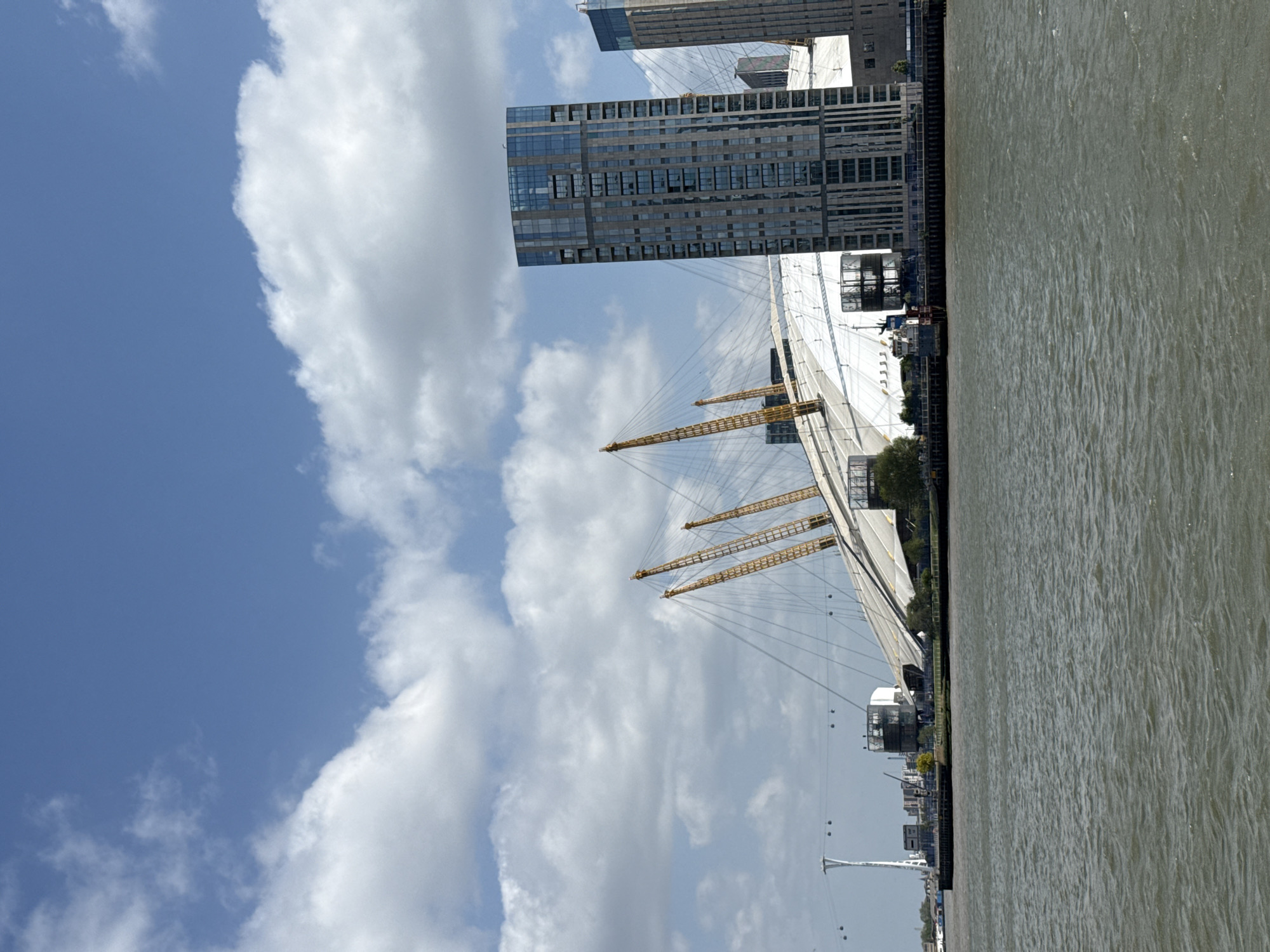 The image captures a bright, sunny daytime scene of the London waterfront, featuring iconic landmarks along the River Thames. The prominent subject in the midground is the O2 Arena, characterized by its distinctive white dome structure, which is supported by several tall, yellow mast-like pillars and a web of cables. Behind the arena, a cluster of modern, tall buildings with reflective glass facades rises against the skyline, indicating a developed urban environment.
To the right, a wide expanse of the River Thames flows, its surface showing ripples under the daylight. The riverbank next to the O2 Arena is lined with further urban infrastructure and some scattered greenery. In the far left of the frame, a tall white mast with suspension cables, identifiable as part of the IFS Cloud Cable Car (Emirates Air Line), is partially visible, further establishing the location as the Greenwich Peninsula in London.
The sky is a clear blue, dotted with scattered white cumulus clouds, suggesting fair weather conditions. The time of day appears to be midday or early afternoon due to the strong, direct sunlight illuminating the scene. No specific human activity or individuals are distinctly visible, and there is no visible text within the image. The overall atmosphere is calm and expansive, showcasing a modern cityscape by the river.