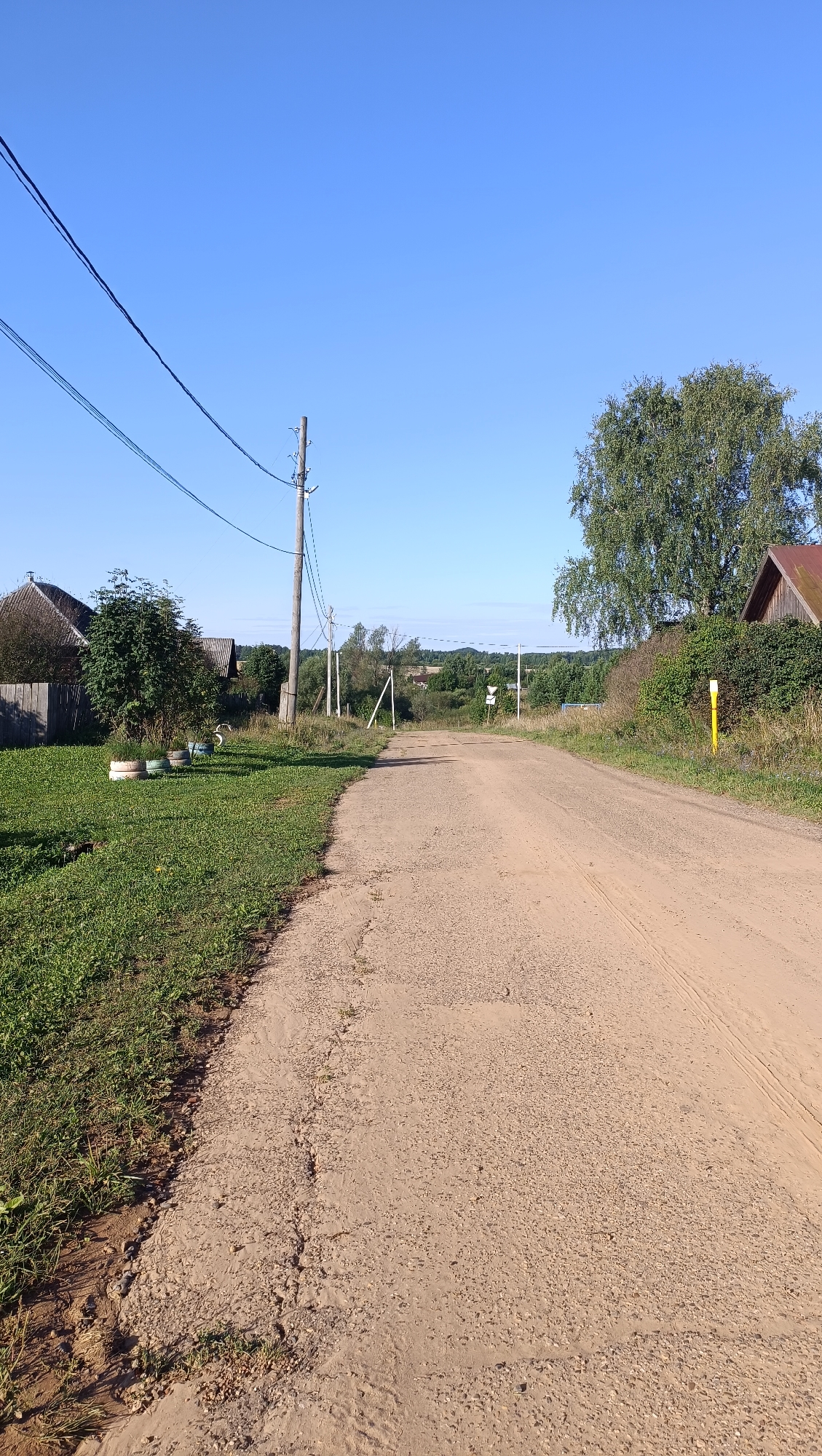 The image depicts a rural road scene on a clear, sunny day. The road, made of gravel and dirt, stretches into the distance, curving slightly to the right. On the left side of the road, a patch of green grass and some scattered tires serve as decoration. Further left, a wooden fence encloses a yard with a house. A utility pole with wires stands prominently near the road. On the right side, a large tree and a section of a building with a reddish roof are visible. The background shows a line of trees and a gently rolling landscape under a bright blue sky. The scene is devoid of any people or active events. The location is identified as Karsha, Russia.