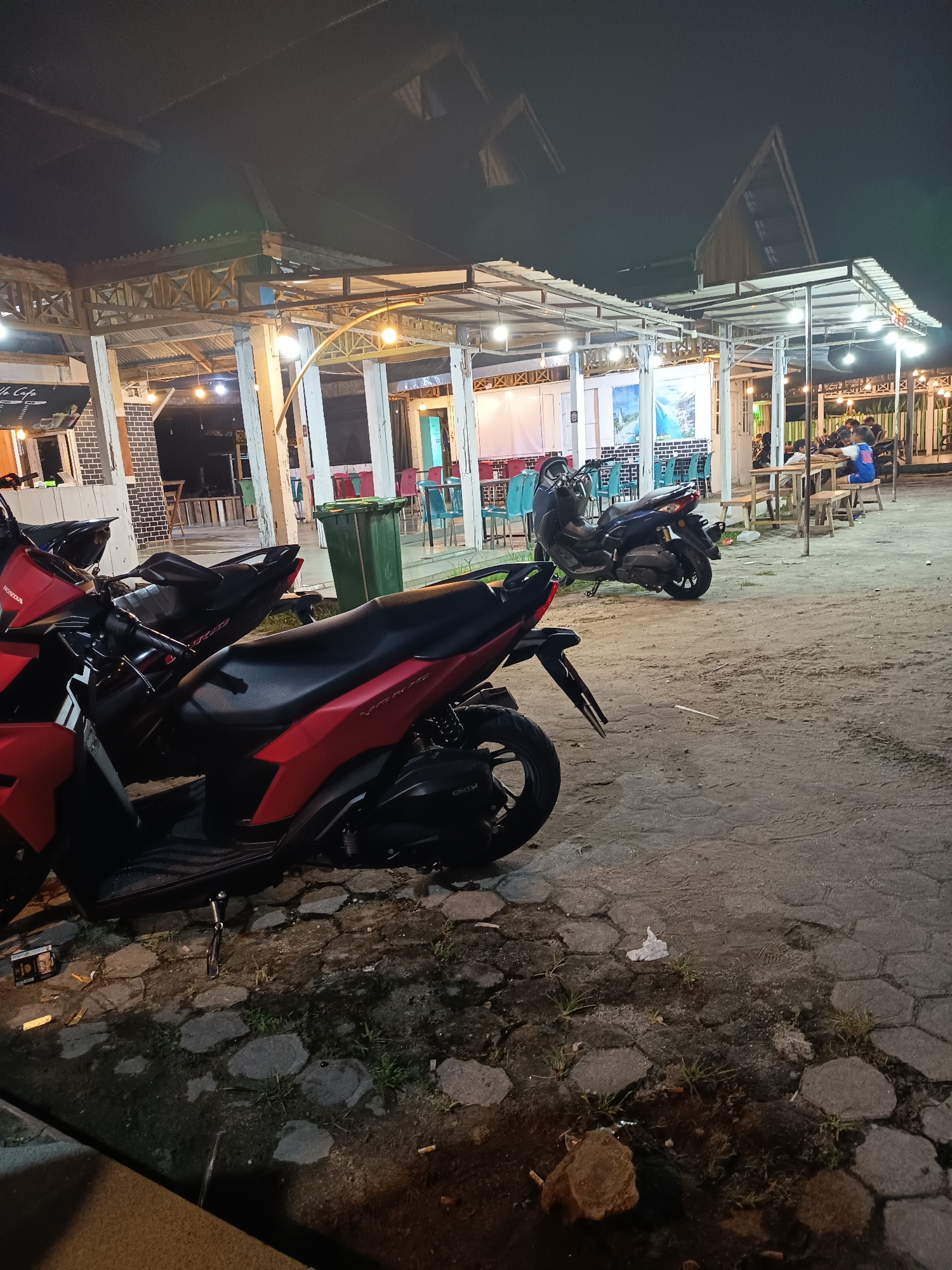 A nighttime scene depicts an outdoor seating area, possibly a cafe or restaurant, in Singkil, Indonesia. Several motorcycles are parked in the foreground, with the closest being a red and black scooter. In the background, people are seated at tables under a covered structure illuminated by artificial lights. The ground is a mix of pavement and sand, with some scattered debris. The overall atmosphere is casual and relaxed.