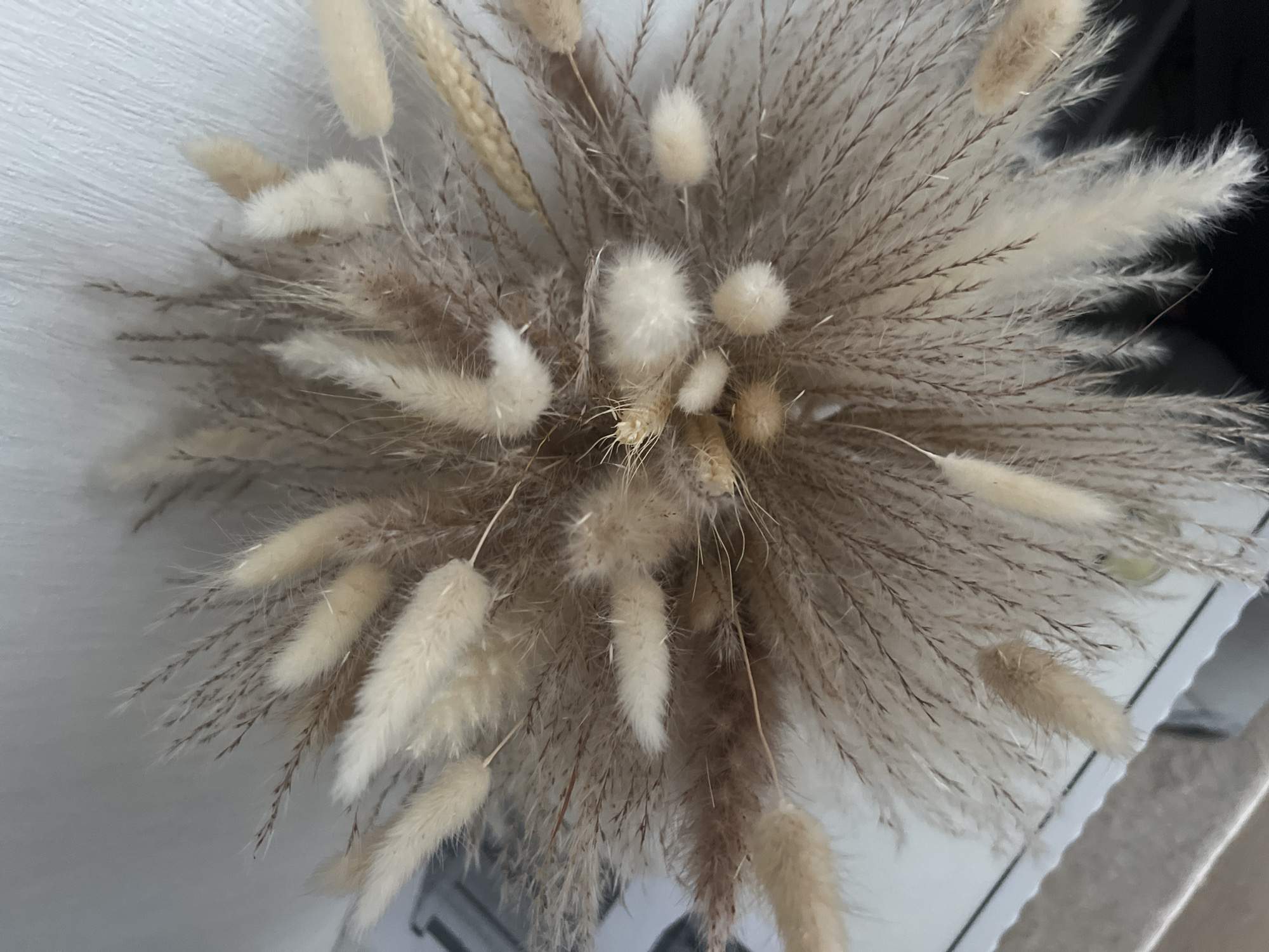 The image is an overhead shot of a dried floral arrangement, likely pampas grass and bunny tails, in a neutral palette of creams and browns. The arrangement is dense, with feathery stalks radiating outwards. The background consists of a white, textured surface, possibly a wall, and a reflective surface below, suggesting a table or shelf. There are no people or animals visible. The scene appears to be an indoor still life, with no specific geographical markers to indicate its location in Votkinsk, Russia.