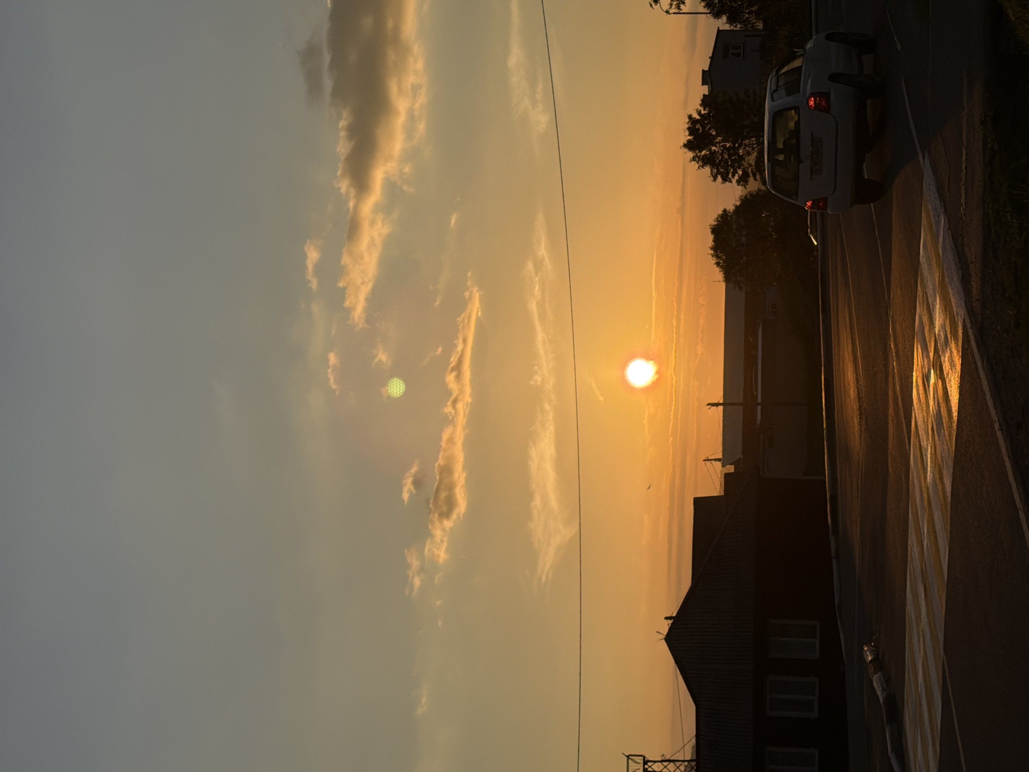 A white SUV drives on a wet asphalt road during sunset. The sun is a bright orange orb, low in the sky, casting a golden glow on the clouds and reflecting on the road. The sky transitions from a pale blue at the top to a warm orange near the horizon. Silhouettes of buildings, trees, and a transmission tower are visible in the background. A pedestrian crossing with yellow and white stripes is prominent in the foreground, showing reflections of the sunset. The scene appears to be in a residential or semi-industrial area, possibly on Sakhalin Island, Russia, given the mention of Yuzhno-Kurilsk, which is part of the Kuril Islands, a Russian archipelago. There are no people visible in the frame.