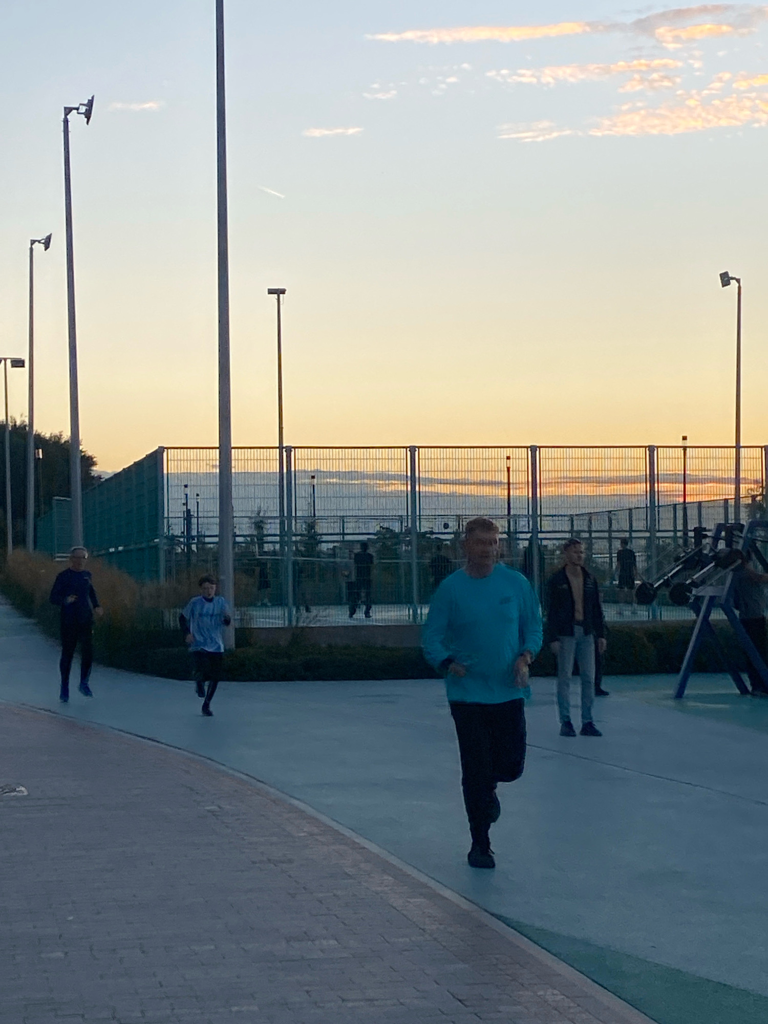 The image depicts an outdoor scene at sunset, possibly in an urban park or recreational area. In the foreground, a man in a teal long-sleeved shirt and black pants is jogging towards the viewer. Behind him, several other individuals are also engaged in physical activity. To the left, two people in dark athletic wear are running away from the camera. Further in the background, beyond a fence and some vegetation, other figures are visible, suggesting a public space with amenities like sports courts or exercise equipment, which are partially discernible through the fence. The sky is a gradient of orange, yellow, and blue, indicative of dusk. Several tall lampposts are scattered throughout the scene, with one prominent one on the left side of the frame. The ground consists of a paved pathway in the foreground and a blue-colored surface further back. There is no specific indication within the image to place it in the city of Perm, Russia.