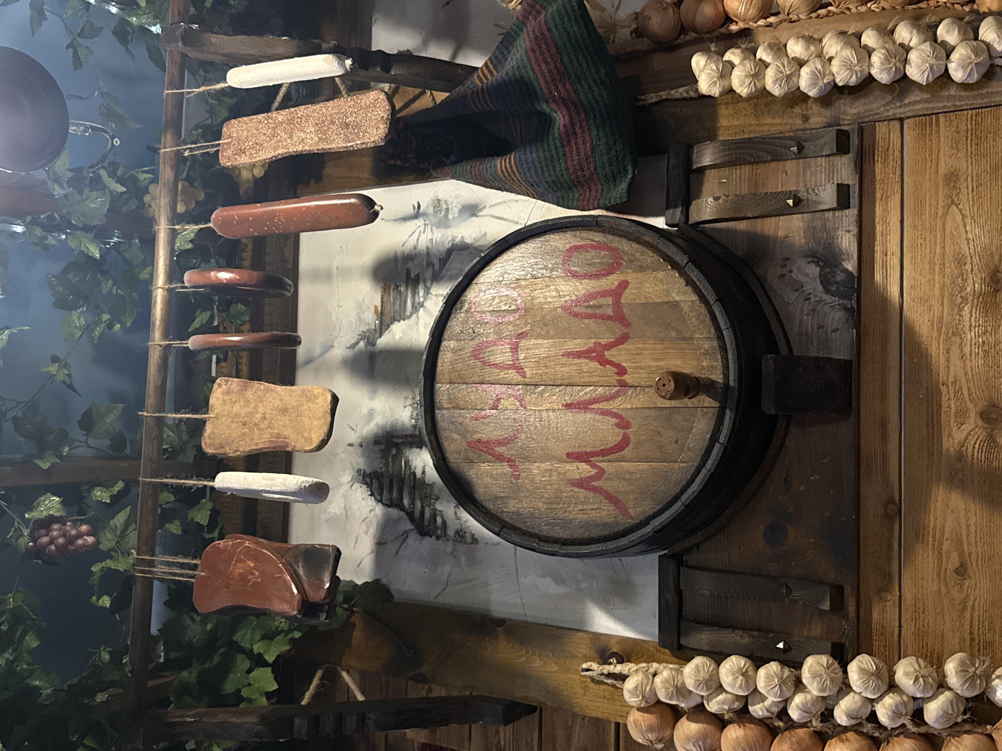 The image depicts a rustic interior scene with a distinct European or old-world aesthetic. The primary focus is a wooden barrel, possibly a wine or liquor barrel, mounted on a wooden wall. Red lettering is painted across the barrel's surface. The wall is adorned with strings of garlic and onions, adding to the old-world atmosphere.
Hanging to the left of the barrel is a display of various meats and sausages, strung up and suspended from a wooden frame. Faux vines and grapes enhance the setting, suggesting a cellar or tavern-like environment. The overall lighting is dim, accentuating the rustic and possibly historical theme.
There are no people visible in the frame, and no specific actions or interactions are depicted. The location is not directly evident from the image, and it is impossible to confirm the city of Des Plaines, United States of America, as the location based solely on this visual information.
