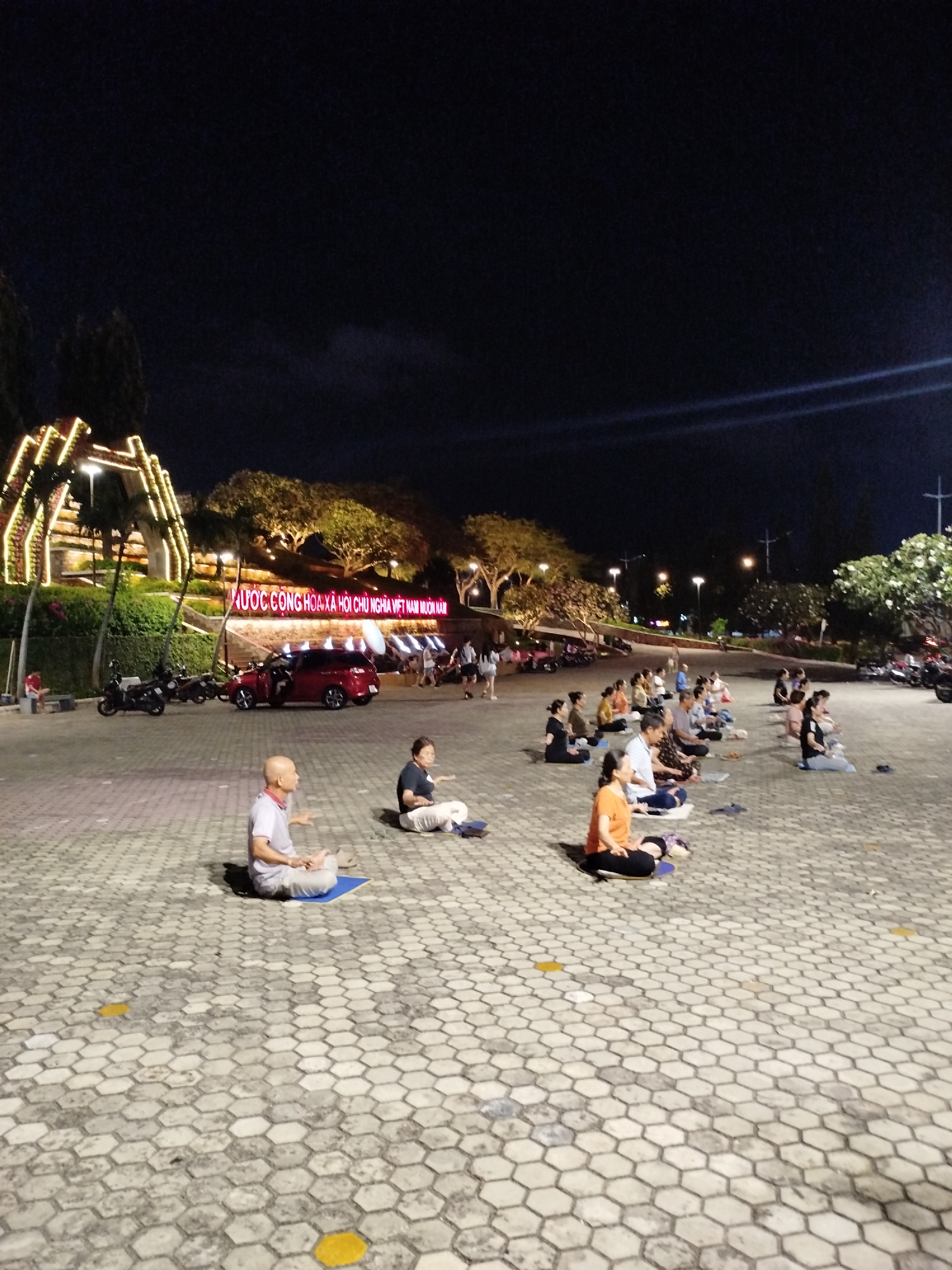 In Ho Chi Minh City, Vietnam, a group of people are seated on a hexagonal-paved surface in an open-air parking lot. They are engaged in an activity, possibly meditation or exercise, as evidenced by their posture and arrangement. A man is seated in a meditative pose on a blue mat. Behind the seated group are parked motorcycles and a red car. In the background, there's a lit-up archway structure. Trees and streetlights illuminate the area. A sign in Vietnamese is visible. The setting is at night. People are walking around in the background.