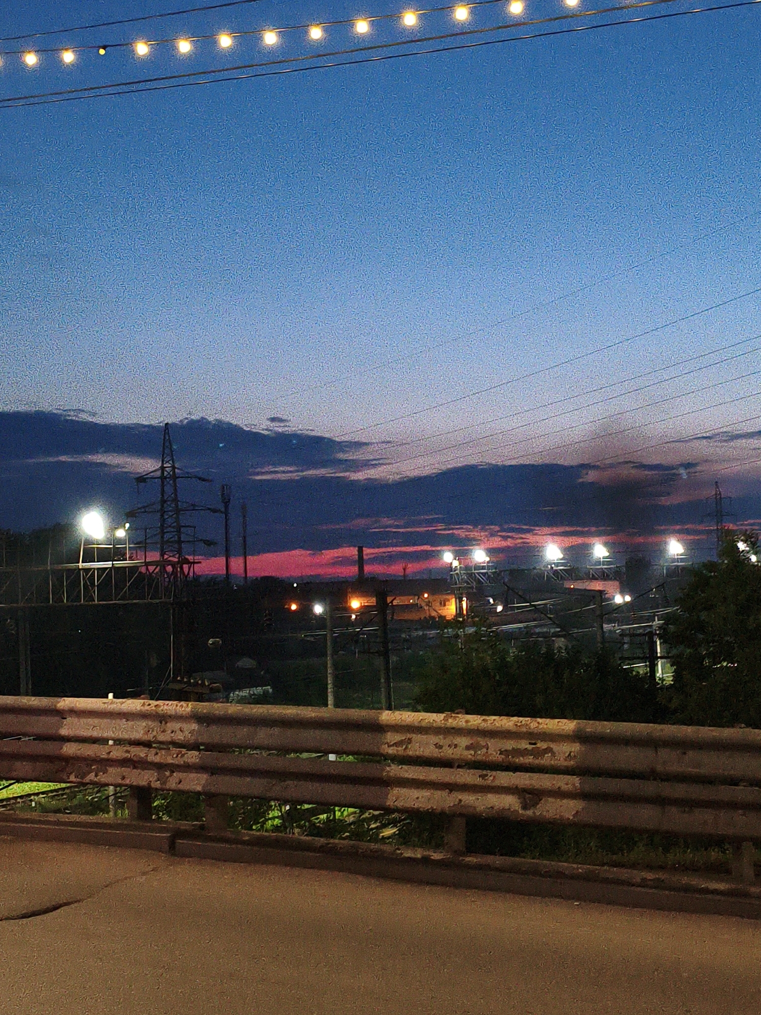 The image depicts a twilight scene, likely taken from an elevated position such as a bridge or overpass, overlooking an industrial area at dusk. The sky is a gradient of deep blue, transitioning to streaks of vibrant pink and orange near the horizon, indicating the sunset or afterglow. Several power lines stretch across the upper portion of the frame, with a string of festive lights hanging above them.
In the middle ground, a prominent electrical pylon stands tall, silhouetted against the colorful sky. Beyond it, a series of industrial structures are visible, some with illuminated lights, suggesting ongoing activity. Trees and foliage are sparsely distributed, with dense green areas on the right side of the image.
In the foreground, a weathered metal guardrail runs horizontally across the frame. It appears to be made of multiple horizontal beams, showing signs of rust and wear. The surface in front of the guardrail appears to be asphalt, with some cracks visible.
The scene is devoid of any human figures or discernible activity. The overall impression is one of urban industrial landscape at the transition from day to night, with a touch of celebratory lighting from the string of bulbs. Given the request to locate this in Orenburg, Russia, it would represent a view of an industrial zone within or on the outskirts of the city during twilight.
