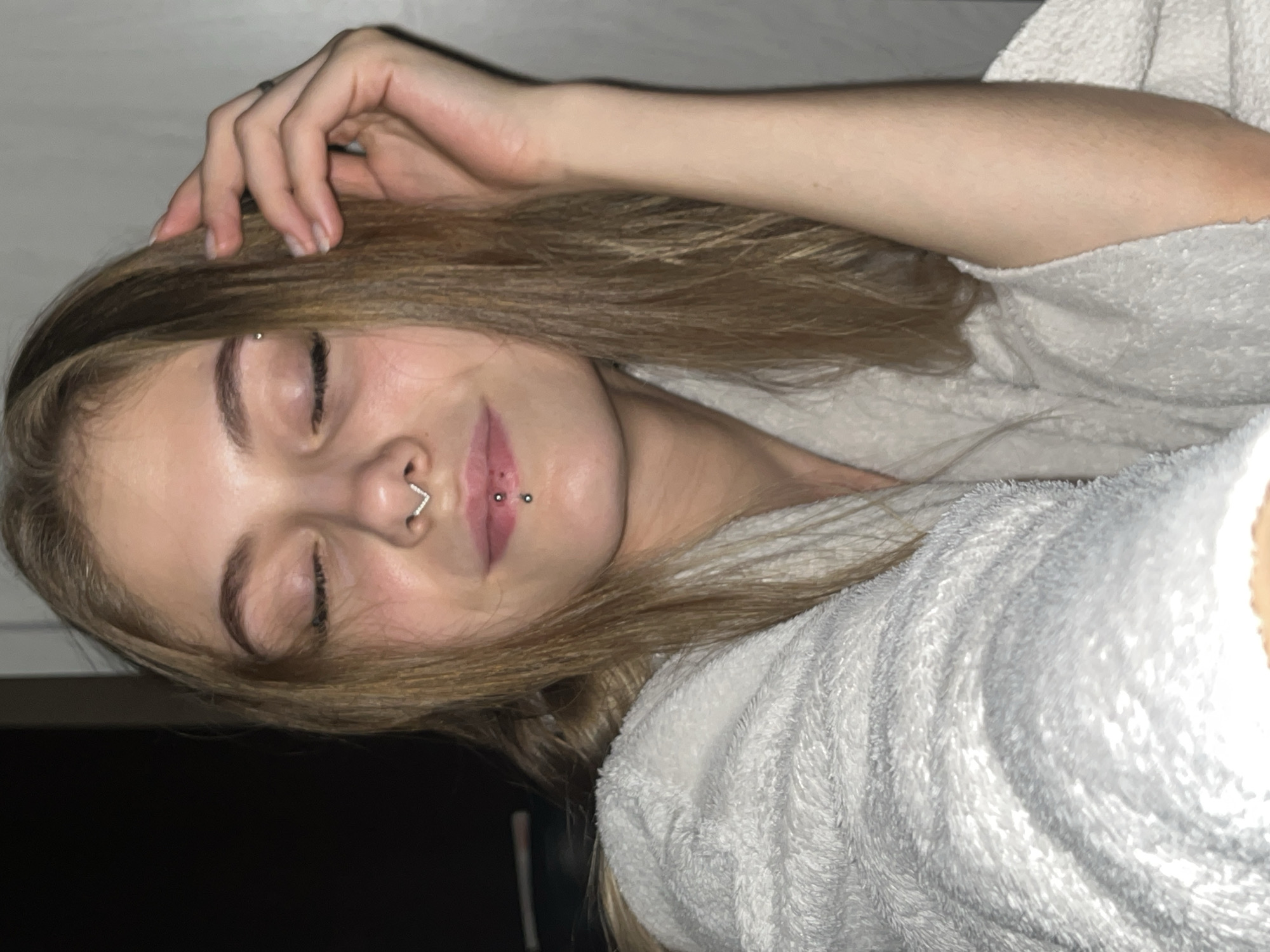 A woman lies on her side wearing a white robe. Her eyes are closed, and her right hand is touching her forehead. She has blonde hair and multiple facial piercings. The background is dark. The location is unknown and cannot be determined from the image.