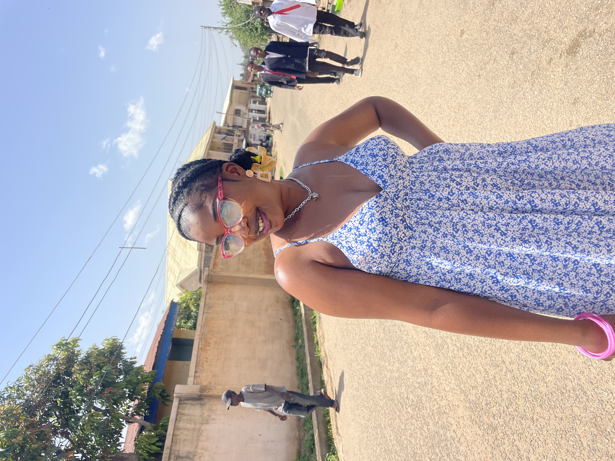 The media file shows a young Black woman with braided hair and pink-rimmed glasses, smiling at the camera. She is wearing a blue and white floral print dress and a silver necklace. Her right arm is bent at the elbow, with her hand on her hip, and she is wearing a pink bracelet.
In the background, several men are walking away from the camera, dressed in suits and ties, suggesting they might be attending a formal event. The scene appears to be a street in Jos, Nigeria. The time of day is clearly daytime, indicated by the bright sunlight and clear blue sky with some scattered clouds. The weather seems to be warm and sunny.
Further in the background, there are buildings and trees, typical of an urban or semi-urban environment. A power line runs overhead. Another person, a man wearing a striped shirt and a cap, is walking towards the camera in the lower left portion of the image. The overall mood is cheerful, with the woman's smile conveying happiness. No visible text is present in the image.