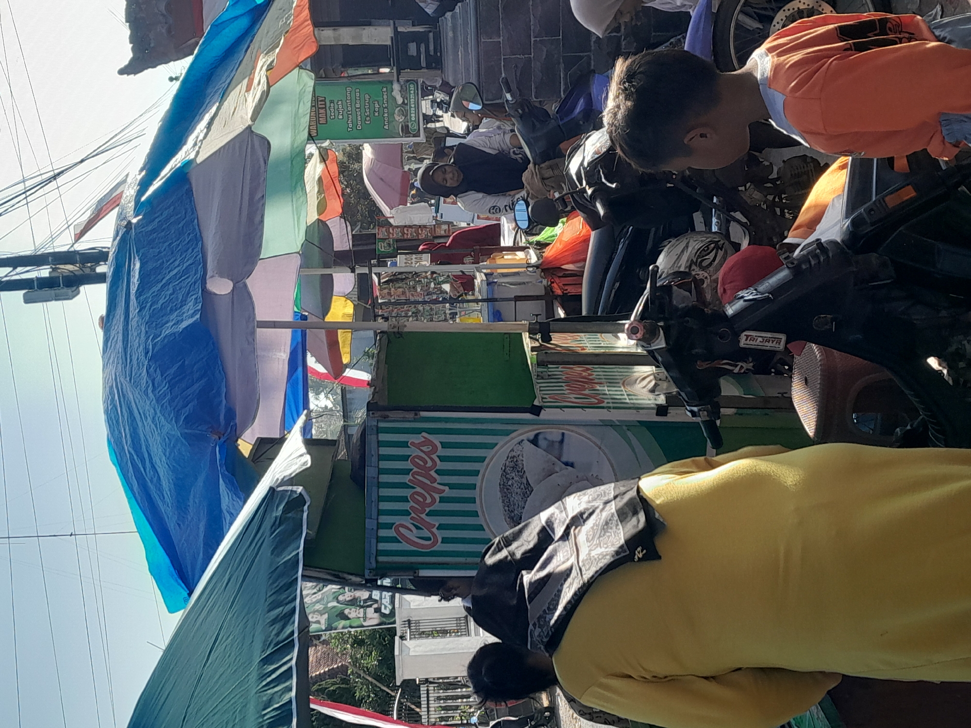 In the city of Bulurejo, Indonesia, a street scene unfolds under a bright, clear sky, with overhead power lines and a tall utility pole visible. Several colorful umbrellas, some plastic and some fabric, are pitched, providing shade for vendors and customers. A green and white striped cart, labeled "Crepes," displays a circular graphic of a crepe. To the right, another stall features a green sign with white lettering listing various food items and a phone number.
In the foreground, a person in a bright yellow outfit stands with their back to the camera, a patterned cloth draped over their shoulders. To their left, a dark-haired child in an orange shirt and light blue shorts looks to the right. Various motorcycles are parked around the stalls. A woman wearing a black hijab and a white shirt is seated on a motorcycle. Other individuals, including a person in a red hooded garment and a woman in a light-colored hijab, are visible in the background, amidst the bustling street market.