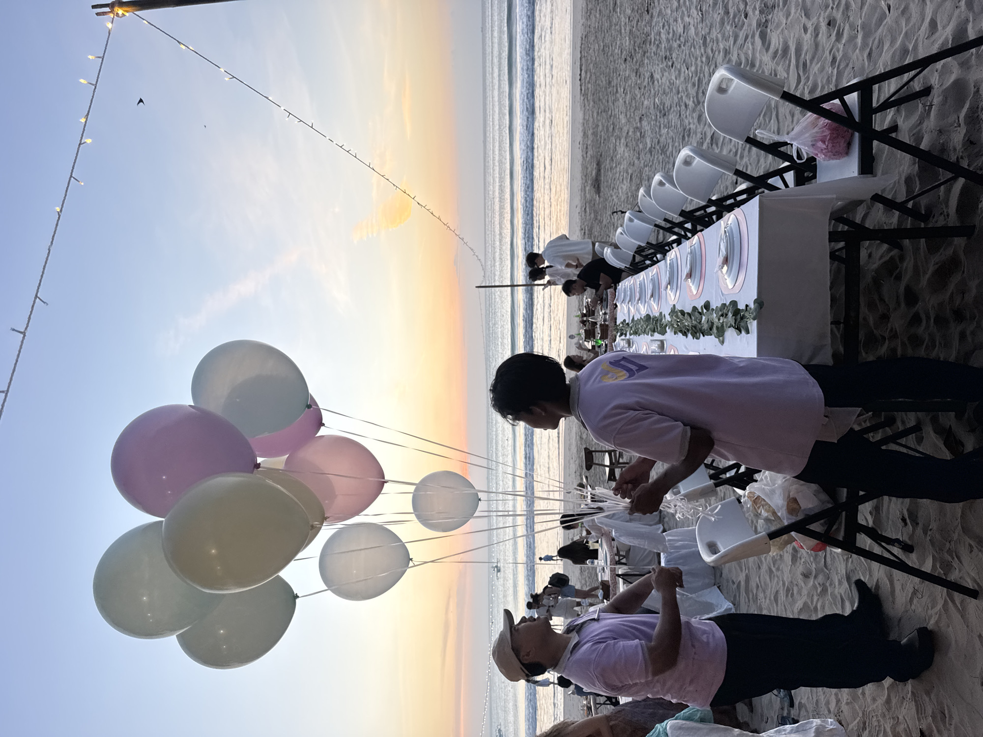 At sunset on the beach in Kuta Selatan, Indonesia, two men in lavender shirts and dark pants are preparing a set of balloons for an event. One man wears a tan baseball cap and holds the balloons. The other man, with short dark hair, holds the strings of the balloons. In the background, a long table is set for dining on the sand, with white chairs arranged on either side. The ocean is visible behind the table, with gentle waves and a few boats in the distance. String lights are strung across poles above the beach, and a bird is flying in the sky. The overall scene suggests an outdoor celebration or gathering.