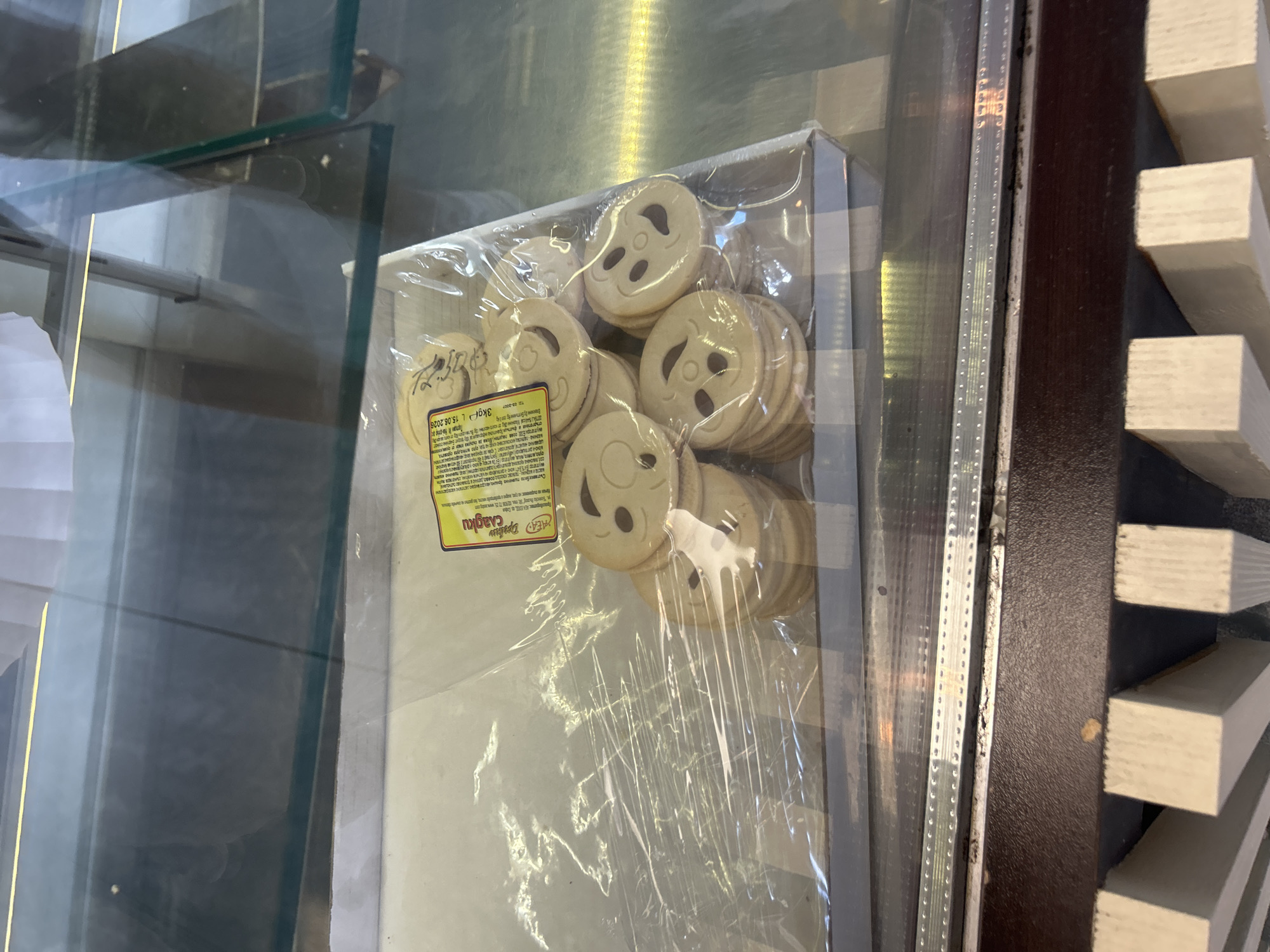 A close-up, eye-level shot shows a plastic-wrapped package of round cookies with smiley face imprints. The cookies are stacked and appear to be a light brown color. A yellow label with text in Cyrillic and a weight of "3kg" is visible on the packaging. The package is resting inside a white cardboard box, which is on display behind a glass counter. The counter and its reflection, along with a wooden structure below, suggest the setting is a retail establishment, possibly a bakery or confectionery shop, in the city of Varna, Bulgaria. The lighting appears to be artificial, creating reflections on the glass and plastic.