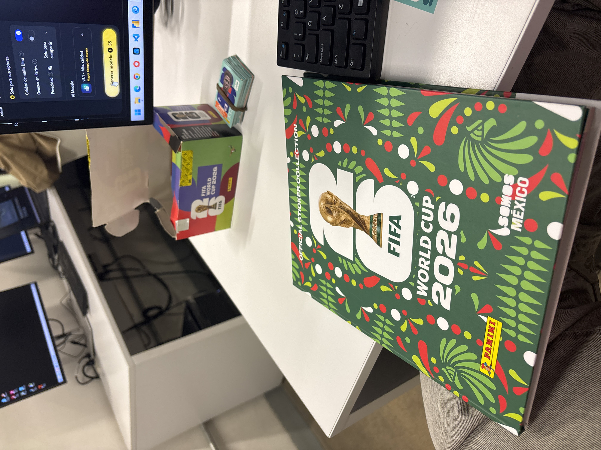 The image shows a desk with a monitor, keyboard, FIFA World Cup 2026 Panini sticker album, a box of FIFA World Cup 2026 stickers, and a stack of individual stickers. The Panini album is green with red and white accents and the words "Somos Mexico." The box of stickers is red and green and has the FIFA World Cup 2026 logo. The monitor displays a software interface with settings for "Calidad de malla Ultra" and a "Generar modelo" button. The desk is white and appears to be in an office setting. The location is likely Mexico City, Mexico due to the "Somos Mexico" text on the album.