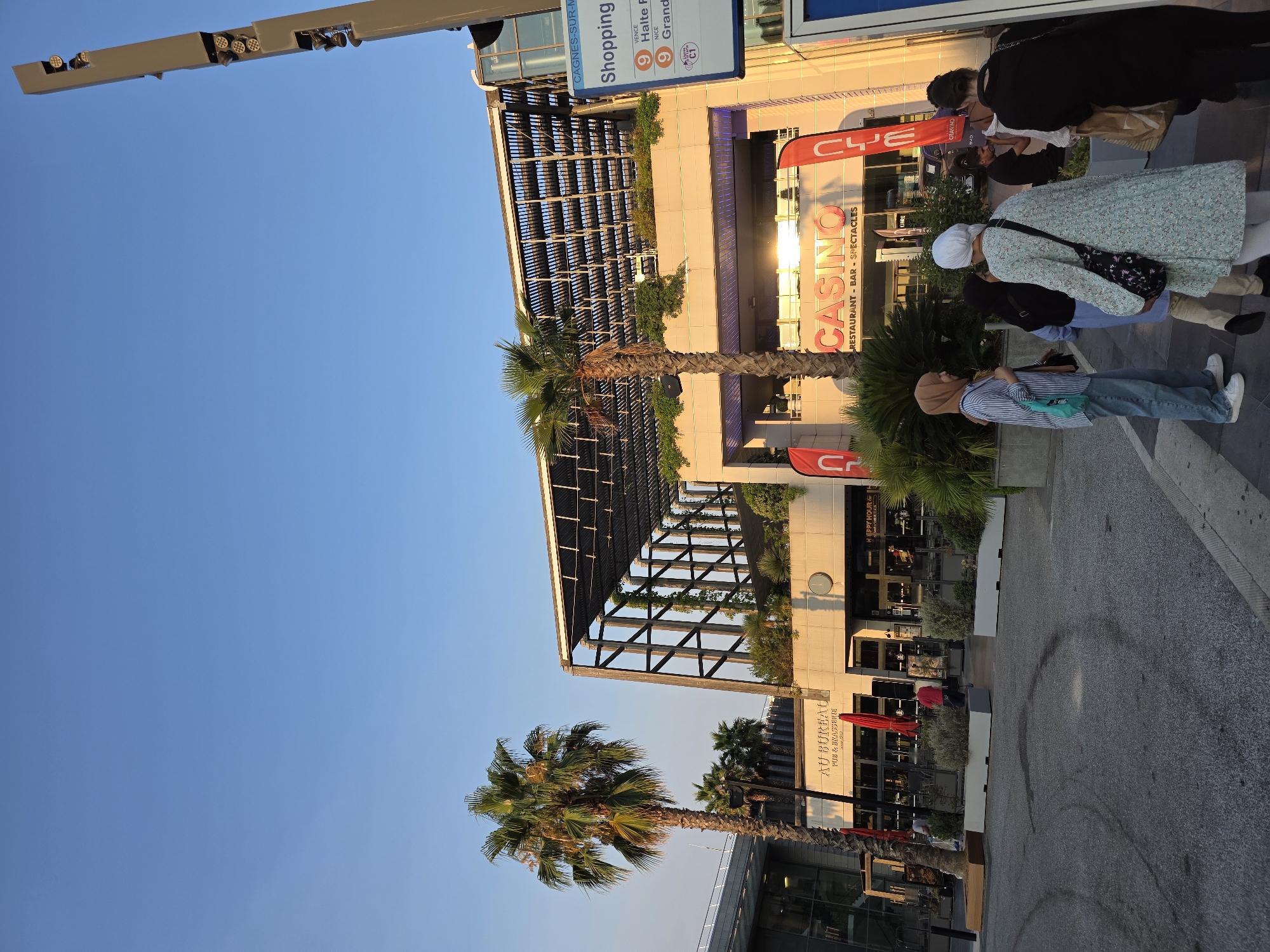 In Cagnes-sur-Mer, France, a bright, sunny day illuminates a modern architectural complex featuring a casino and shopping area. A tall, slender tower with multiple spotlights is positioned on the right side of the frame. Palm trees, one prominently in the foreground, add a tropical touch to the scene. The building itself has a sleek, contemporary design with a slatted roof structure. Signs indicate "CASINO RESTAURANT - BAR - SPECTACLES" and "AU BUREAU PUB & BRASSERIE". A group of people, including women wearing hijabs, are gathered in front of the establishments. The ground is paved, with some dark tire marks visible. A blue signpost with bus route information for "CAGNES-SUR-MER SHOPPING" is also present.