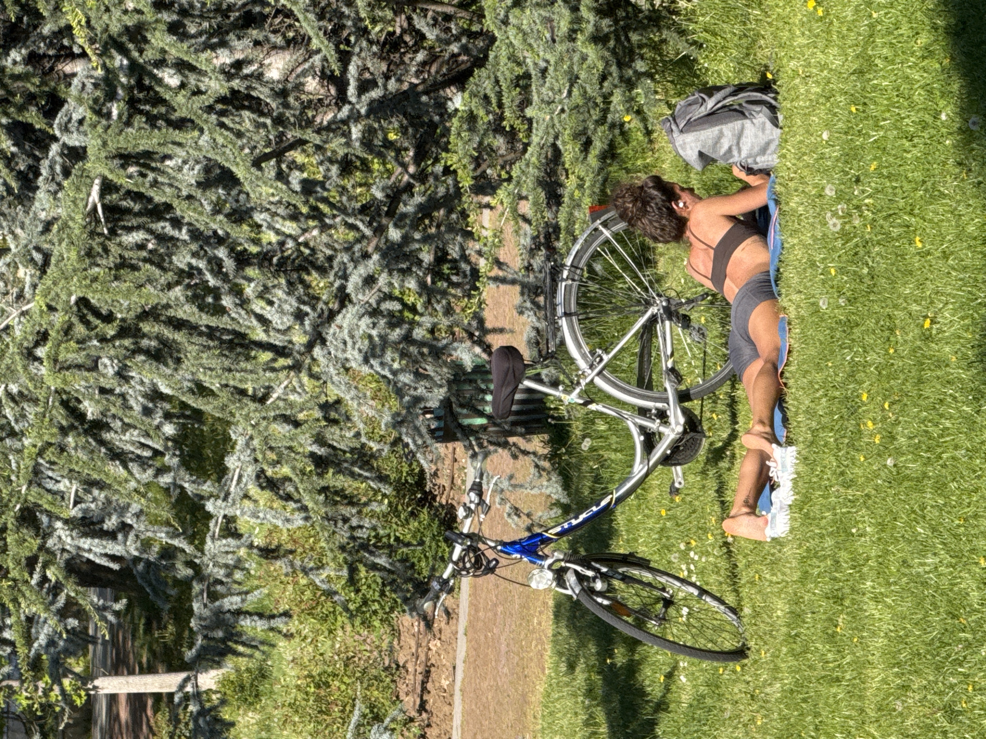 A woman is lying on a blue blanket on the green grass. She is wearing a black sports bra and gray shorts, with white sneakers on her feet. Her bicycle is next to her, and a gray backpack is near her head. There are dandelions scattered in the grass. Behind her is a large green tree with dense foliage. The location is Sofia, Bulgaria.