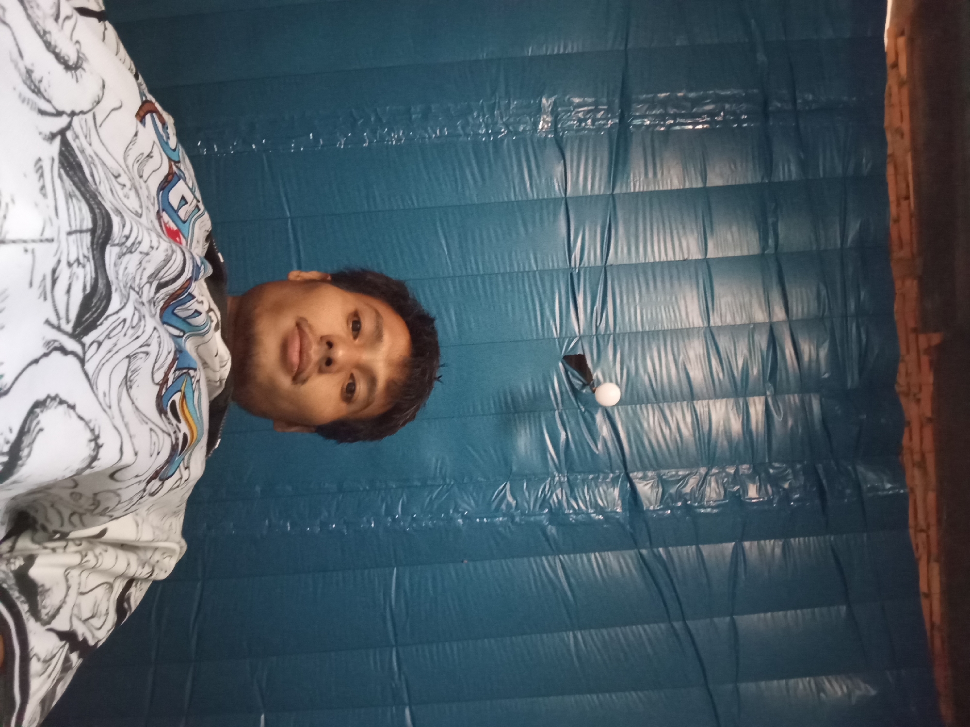 The image, captured at an unknown date and time in Tebo Jaya, Indonesia, depicts a low-angle, close-up, self-taken portrait of a young adult male. The subject is positioned in the center-left of the frame, facing towards the camera with a neutral expression. He has dark hair and is wearing a white long-sleeved garment with a black and colored graphic print. The background is a solid, textured, dark blue surface, possibly a wall or curtain, which appears to be made of padded material with horizontal stitching or seams. A small, spherical, white object is visible in the upper right quadrant of the image, against the blue background. The lighting is dim and appears to be artificial, casting highlights on the textured background and on the subject's face and clothing. The overall scene is indoors, with no visible windows or distinct environmental features to indicate the specific location within Tebo Jaya.