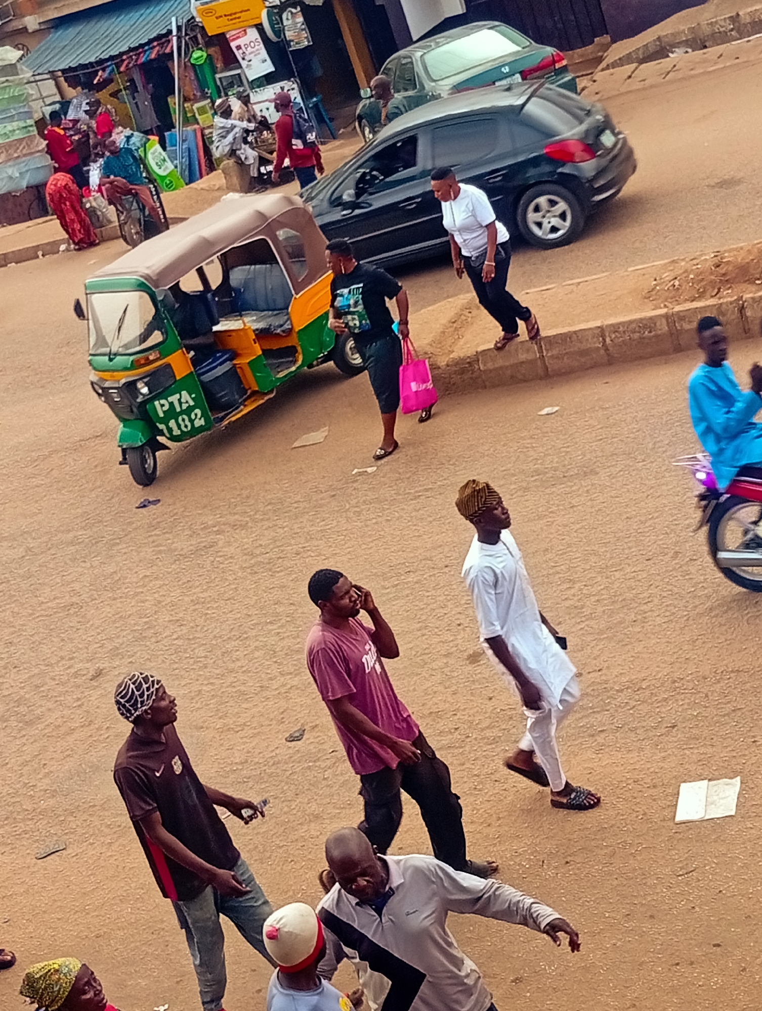 This photo, taken in Bukuru, Nigeria, captures a bustling street scene. In the foreground, four men are walking; one wearing a maroon shirt is on a phone call. In the midground, a woman is stepping off the curb near a dark car, while another woman with a pink bag walks near a green and yellow tricycle (auto-rickshaw) marked with the license plate "PTA-1182". In the background, a shop with mattresses on display is visible, as well as other vehicles and pedestrians on the street. The street is unpaved and has a reddish-brown color. A motorcyclist in blue clothing drives by in the middle ground to the right.