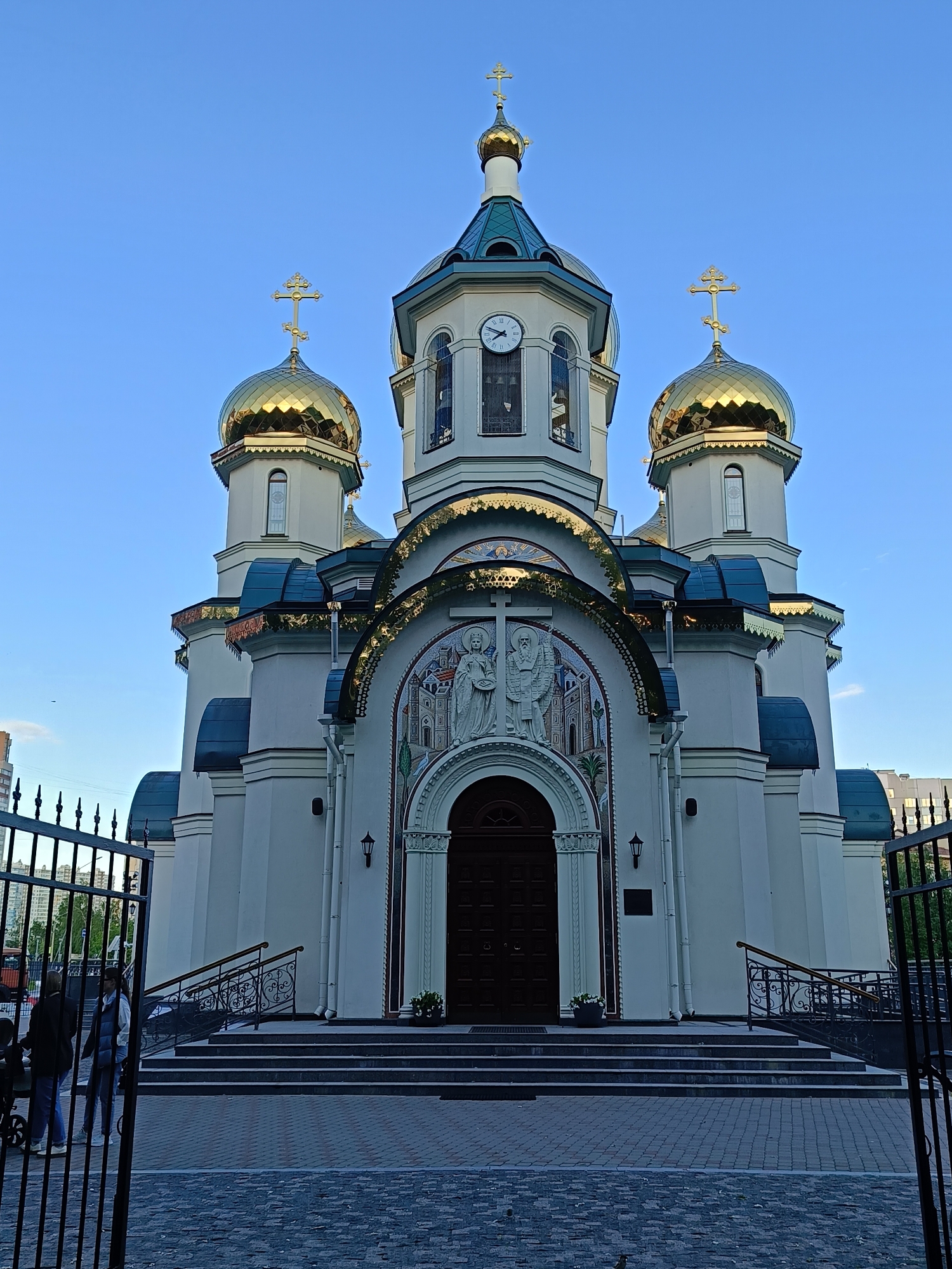 The image depicts a white Orthodox church with golden domes and blue roofs. The central tower features a clock. The church is adorned with mosaics above the entrance. A black wrought-iron fence partially obscures the foreground, and two people, one with a stroller, are visible near the fence. The background shows other buildings, suggesting an urban location. The sky is a clear blue. The location is in the city of Unknown, Unknown.