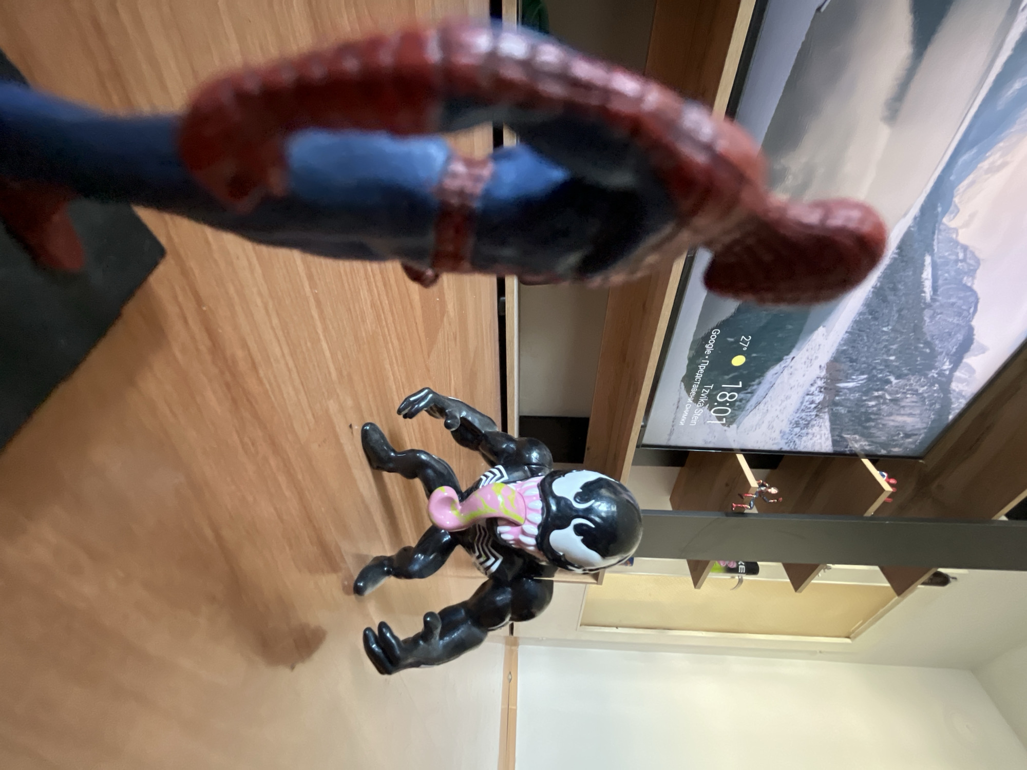The image depicts a domestic indoor scene. In the foreground, two action figures, Spider-Man and Venom, are positioned on a wooden floor. Spider-Man, a red and blue figure, is partially visible on the left, facing away from the camera. Venom, a black figure with a long pink tongue, is on the right, facing Spider-Man. Behind the figures, a large flat-screen television displays a scenic mountain landscape with snow-covered peaks and a body of water. The television screen also shows the time as 18:01 and the temperature as 27 degrees Celsius, with text that appears to be in Bulgarian, possibly indicating location or weather information. To the right of the television, a shelf unit is visible with several items, including small action figures and spray cans. The overall lighting suggests an interior environment, possibly a living room. There is no indication of the city of Shumen, Bulgaria, in the image itself.