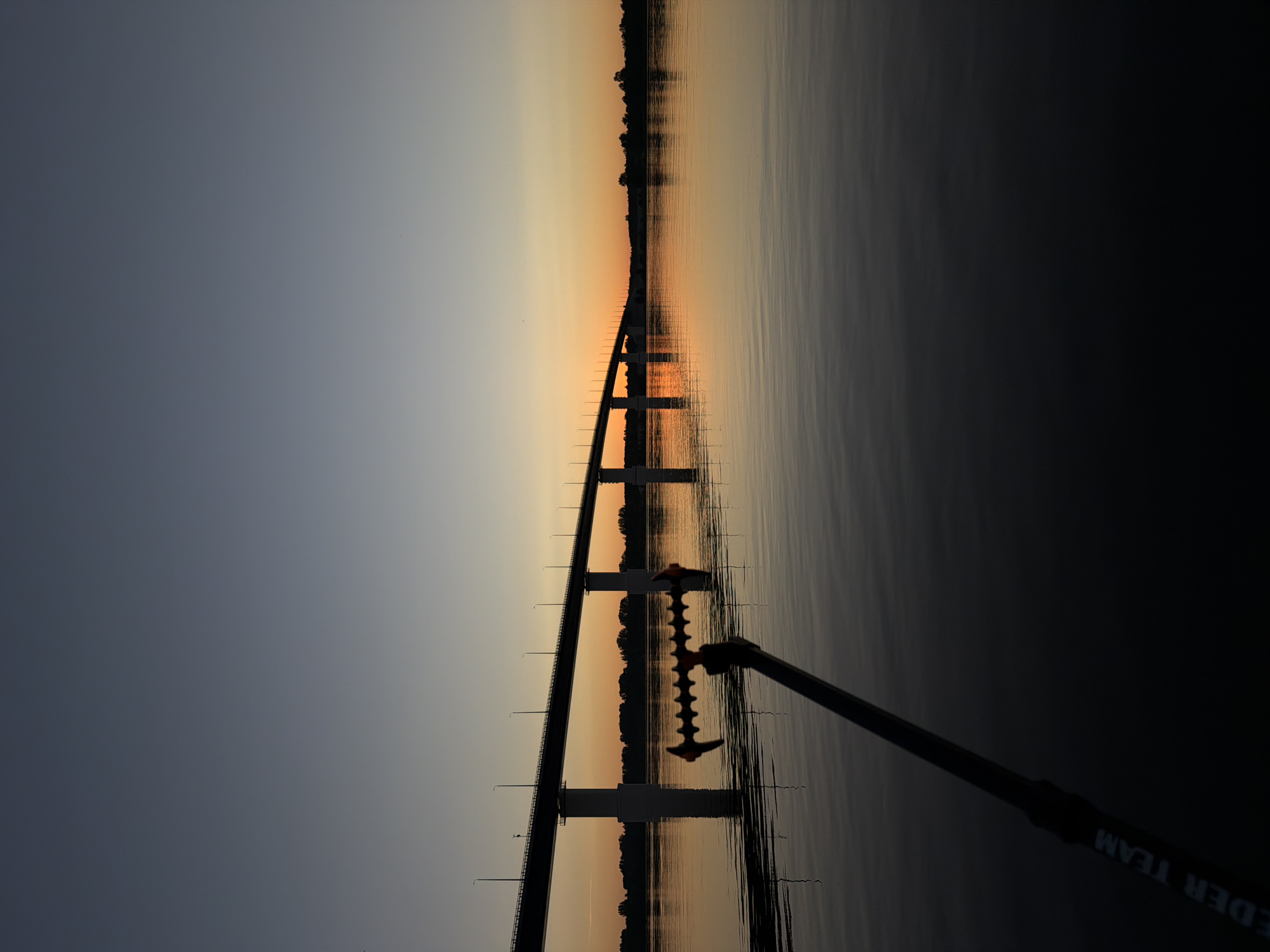A wide shot captures a tranquil river scene at sunset, featuring a long bridge spanning across the water. The sky displays a gradient of colors, from a deep blue at the top to warm orange and yellow hues near the horizon. The bridge, supported by several concrete pillars, has a railing and a series of lampposts running along its length. The water's surface is calm, reflecting the bridge and the colorful sky, creating a mirror-like effect. In the foreground, a portion of a fishing rod with a specialized rest is visible, angled towards the water. The rest has an orange and black design with multiple indentations. The rod itself is dark and has white lettering that reads "FEIDER TEAM". The background shows a silhouetted treeline on the far bank of the river, indicating land. The overall atmosphere is peaceful and serene, suggesting a quiet moment by the water during twilight.