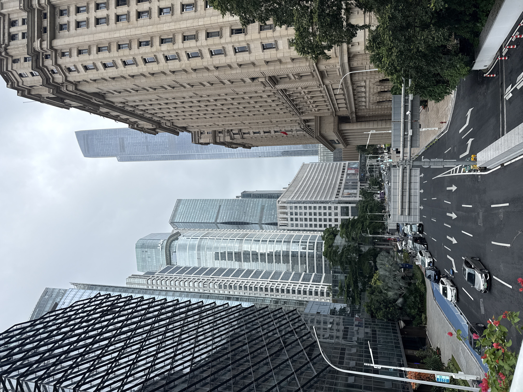 In Tianhe District, China, a street scene unfolds beneath a cloudy sky. The viewpoint is from an elevated position, looking down upon a multi-lane road lined with cars. To the left, a modern glass-faceted skyscraper with a distinct geometric facade dominates the frame. To the right, an older, ornate stone building with numerous windows and classical architectural details stands prominently. In the background, a series of contemporary glass skyscrapers, some with rounded edges, rise into the sky. The road itself has directional arrows painted on its surface. A designated blue lane for cyclists or pedestrians runs along the left side of the road. A few people are visible on the sidewalk near the buildings. Green trees and some flowering plants add a touch of nature to the urban landscape. The overall impression is one of a bustling, modern cityscape where different architectural styles coexist.