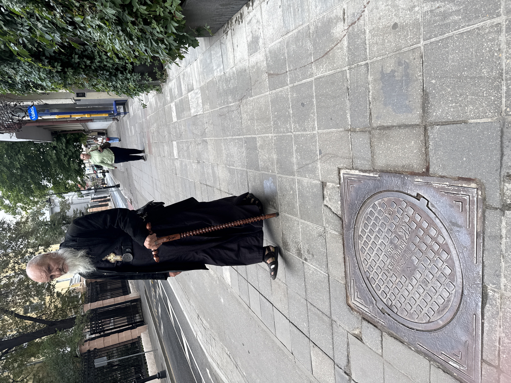 A bald, elderly man with a long white beard, wearing a black robe and sandals, walks along a grey tiled sidewalk in Moscow, Russia. He carries a dark wooden rosary in his right hand. In the background, a woman in a green jacket and black pants walks away from the camera. A building with ivy growing on its facade is to the right, and a street with cars is to the left. A manhole cover is visible in the foreground.