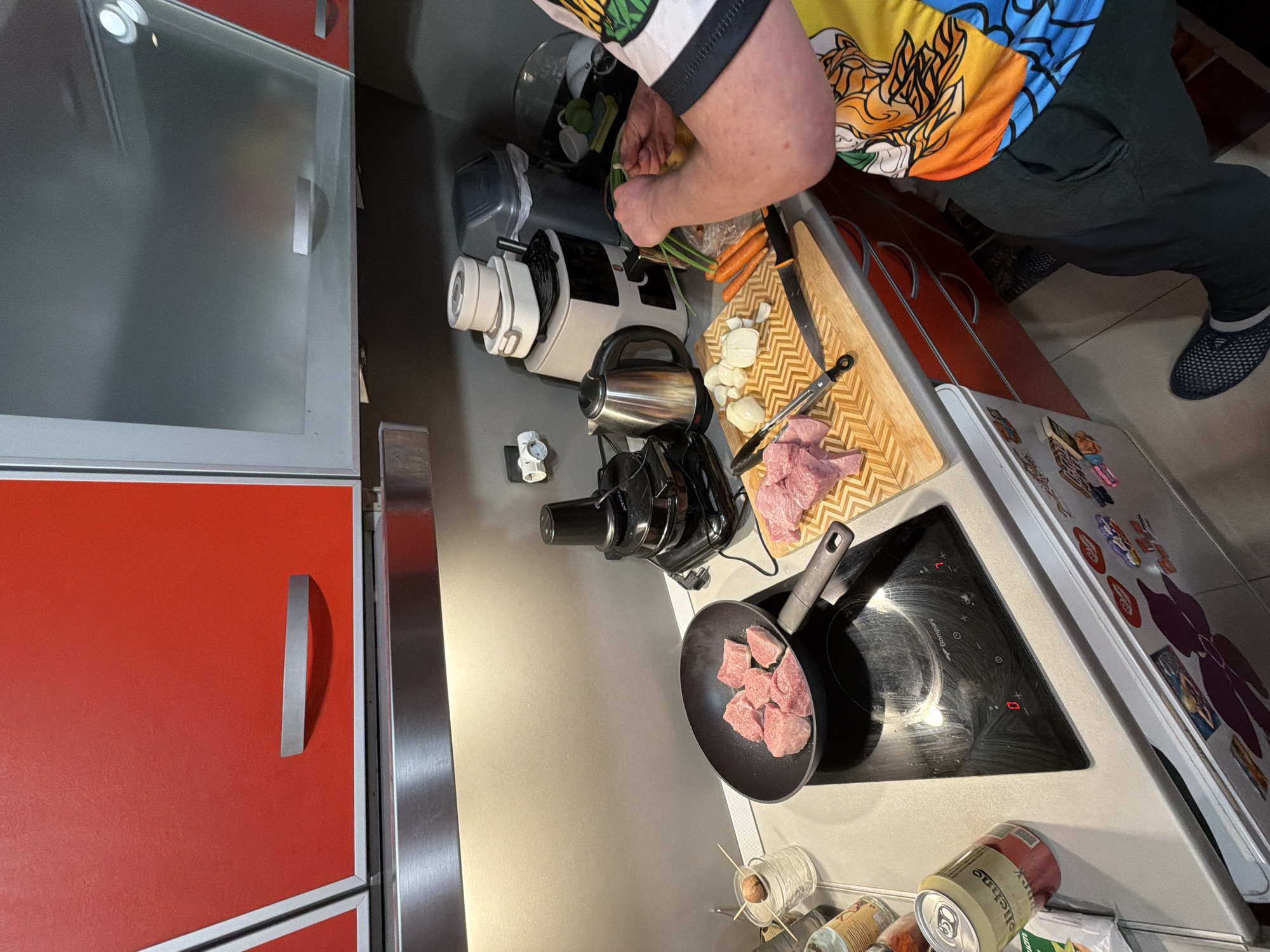 The image depicts a vibrant kitchen scene where a person is engaged in food preparation. The person, whose upper body and legs are visible, is wearing a distinctive colorful short-sleeved shirt with anime-style graphics (resembling Dragon Ball Z characters) and dark trousers. One foot, clad in a blue mesh shoe, rests on a tiled floor.
The central activity revolves around a modern kitchen counter. On an induction cooktop, a black frying pan holds cubes of raw meat, likely pork or beef, ready for cooking. The cooktop itself features illuminated controls and the visible brand name "Bonzanni." Adjacent to it, a wooden cutting board is laden with more raw meat, chopped onions or garlic, and whole carrots, along with green onions that the person appears to be handling or preparing. A kitchen knife and metal tongs are also present on the board.
The counter is well-equipped with various appliances, including a white and black blender or food processor, a stainless steel electric kettle, and a black juicer. Towards the front edge of the counter, several items are placed: clear glass jars, one seemingly sprouting an avocado pit, and various cans and jars, including a green can of "Heineken" and another product labeled "Schneekoppe."
To the left of the scene, modern red and silver kitchen cabinets are visible, with an upper cabinet featuring a glass front that reflects internal lights. To the right, a refrigerator is heavily decorated with numerous colorful magnets and stickers. The lighting in the kitchen appears to be artificial, suggesting an indoor setting where preparation could be happening at any time of day, perhaps evening. The scene conveys a busy yet organized atmosphere of home cooking in what appears to be a contemporary kitchen environment in Varna, Bulgaria.