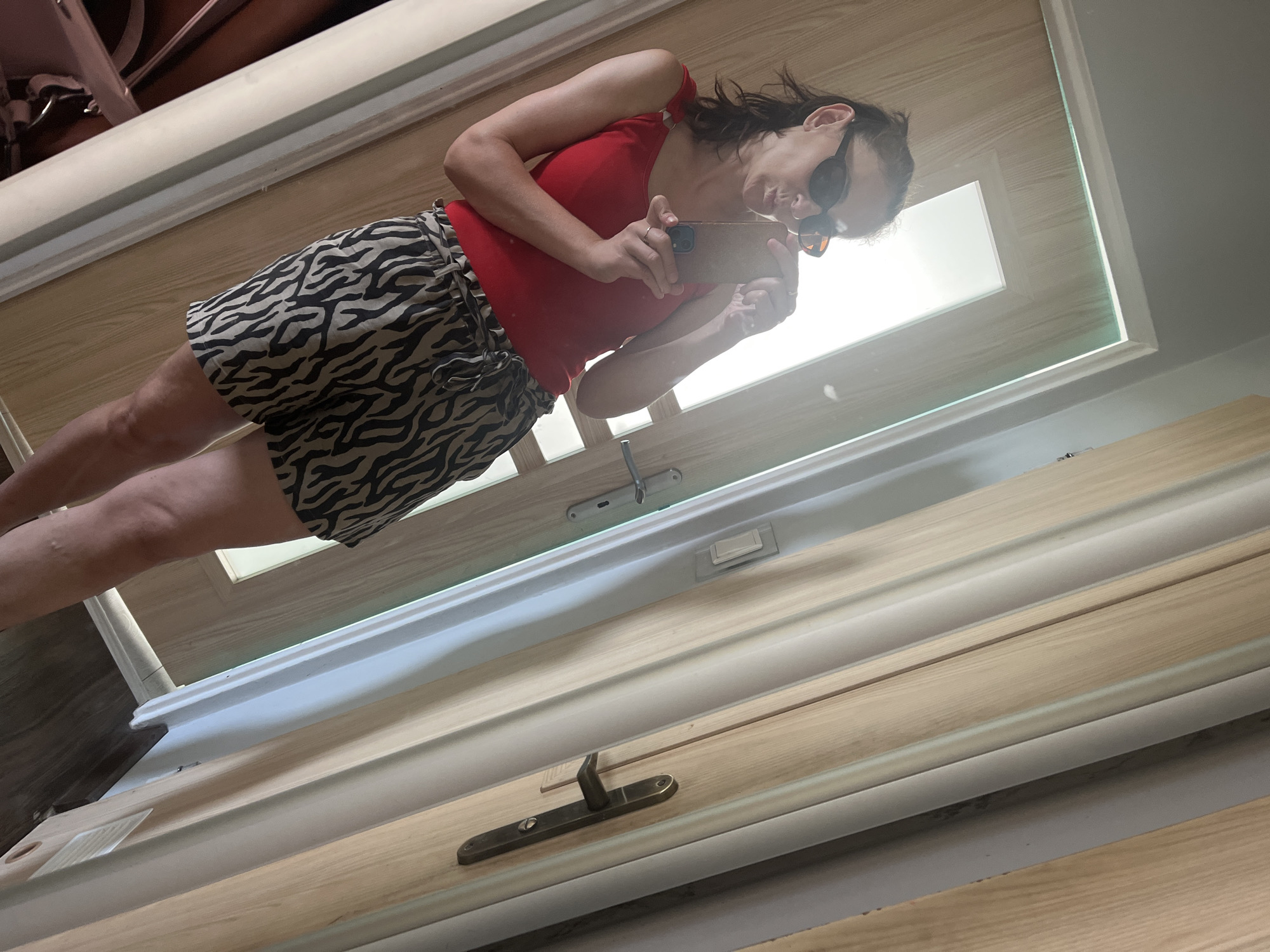 A woman is taking a selfie in a mirror. She is wearing a red tank top and black and white zebra-print shorts. She has on sunglasses and is holding a smartphone in her right hand, pointing it at the mirror. The setting appears to be indoors, possibly a room with a door or large window with a reflective surface. The lighting suggests it is daytime. The woman's expression is unreadable due to the sunglasses. The scene is reflected in what looks like a large pane of glass, likely a door or mirror, with a wooden frame. Wood-paneled walls and trim are visible in the reflection. There is also a door handle visible in the reflection on what seems to be a door.