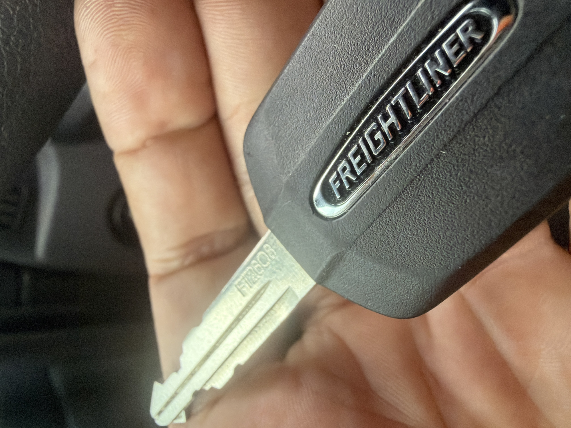 A close-up, eye-level shot shows a person's hand holding a black Freightliner key. The key's blade is silver and has a jagged edge. Embossed on the black plastic part of the key is the word "FREIGHTLINER" in silver lettering. The background is dark and out of focus, with hints of a vehicle interior visible. The image was taken in Murfreesboro, United States of America.