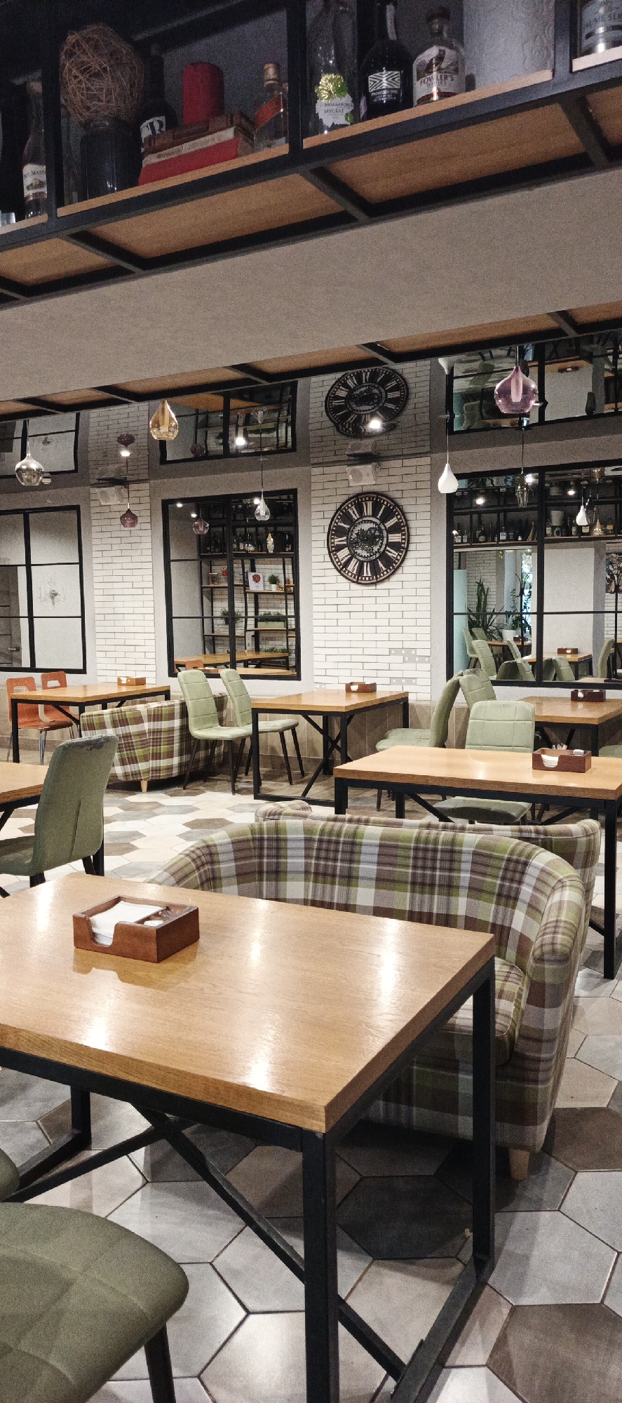 The image depicts the interior of a cafe, possibly located in Votkinsk, Russia. The cafe features a modern rustic aesthetic with wooden tables, metal-framed chairs, and plaid upholstered seating. Shelving units displaying decorative items, bottles, and books are visible on the upper walls. Two large, ornate clocks are prominently displayed on the white brick wall, contributing to the cafe's ambiance. Pendant lights with glass shades hang from the ceiling. The floor is tiled with a geometric pattern of hexagonal and trapezoidal shapes in various shades of grey and beige. The scene is empty of people, suggesting the cafe is either closed or the photograph was taken before or after operating hours. The lighting is subdued, with spotlights highlighting certain areas.