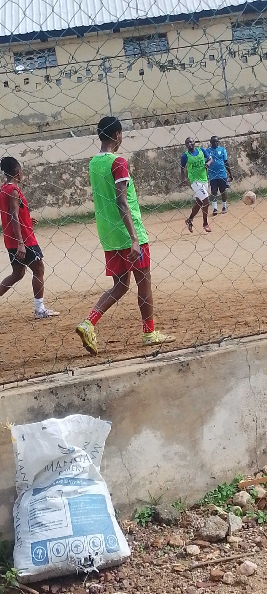 The image depicts a soccer game in progress behind a chain-link fence. Three players are visible in the foreground and middle ground. One player on the left wears a red shirt and black shorts. Another player, closer to the center, is wearing a green training vest over a red shirt and red shorts. A third player is in the middle ground wearing a green training vest over a blue shirt and white shorts. There is also a fourth player in the background wearing a blue shirt. A soccer ball is in the air between the third and fourth players.
In the foreground, at the bottom of the image, there is a bag of MANGAL CEMENT resting on the ground next to a concrete structure. The ground is littered with rocks and sparse vegetation.
Behind the fence, a building with windows is visible. The image appears to be taken outdoors in daylight. Given the presence of the MANGAL CEMENT bag, it's likely the location is Lokoja, Nigeria, where this cement is manufactured.