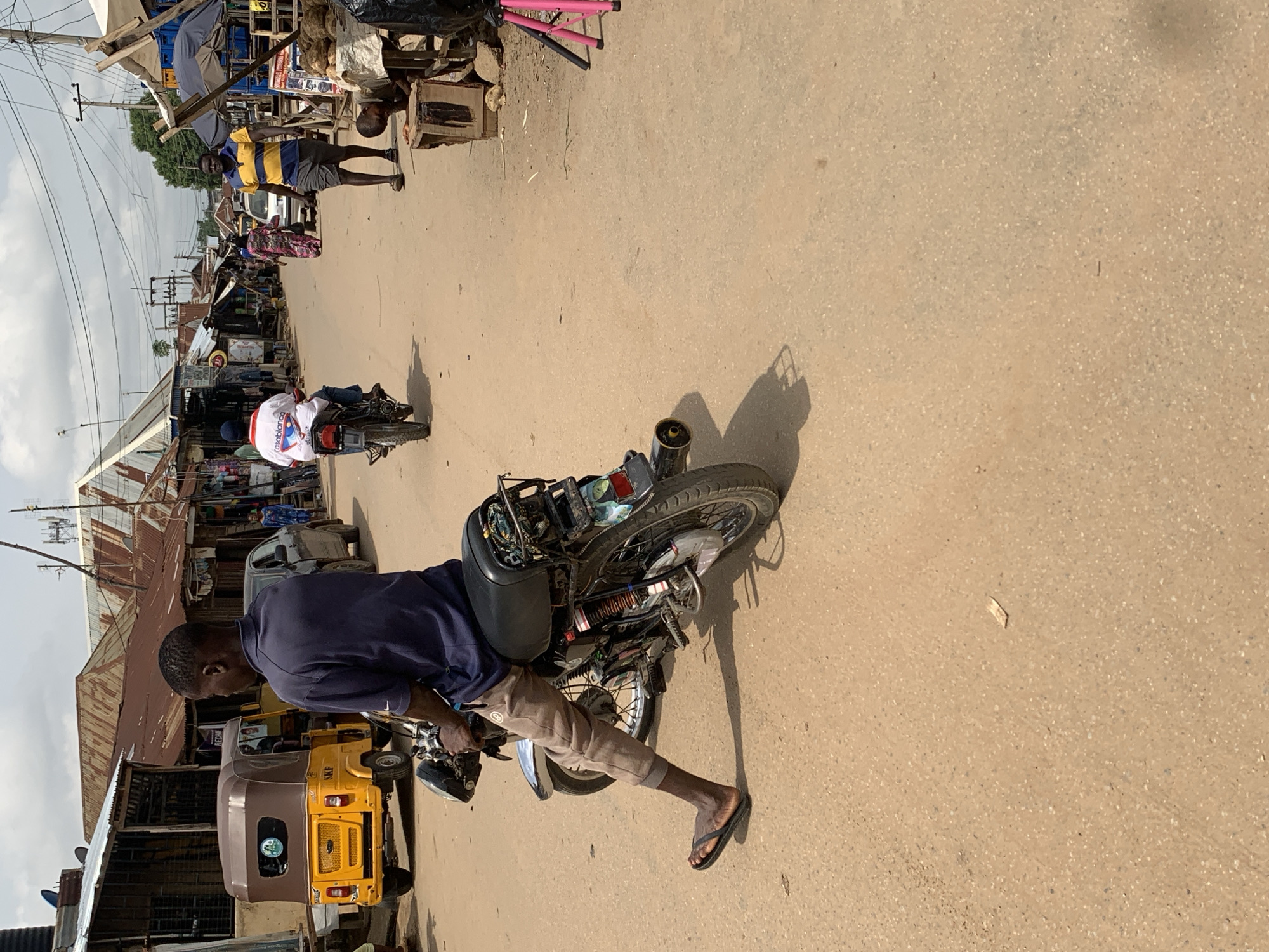A street scene in Abuja, Nigeria, shows a man standing next to a motorcycle, likely his mode of transportation. He is wearing a blue shirt and khaki pants with flip-flops. The motorcycle is heavily customized and loaded. In the background, there are shops, people, and a yellow three-wheeled vehicle (Keke Napep). The road appears to be paved. The lighting suggests it is daytime.