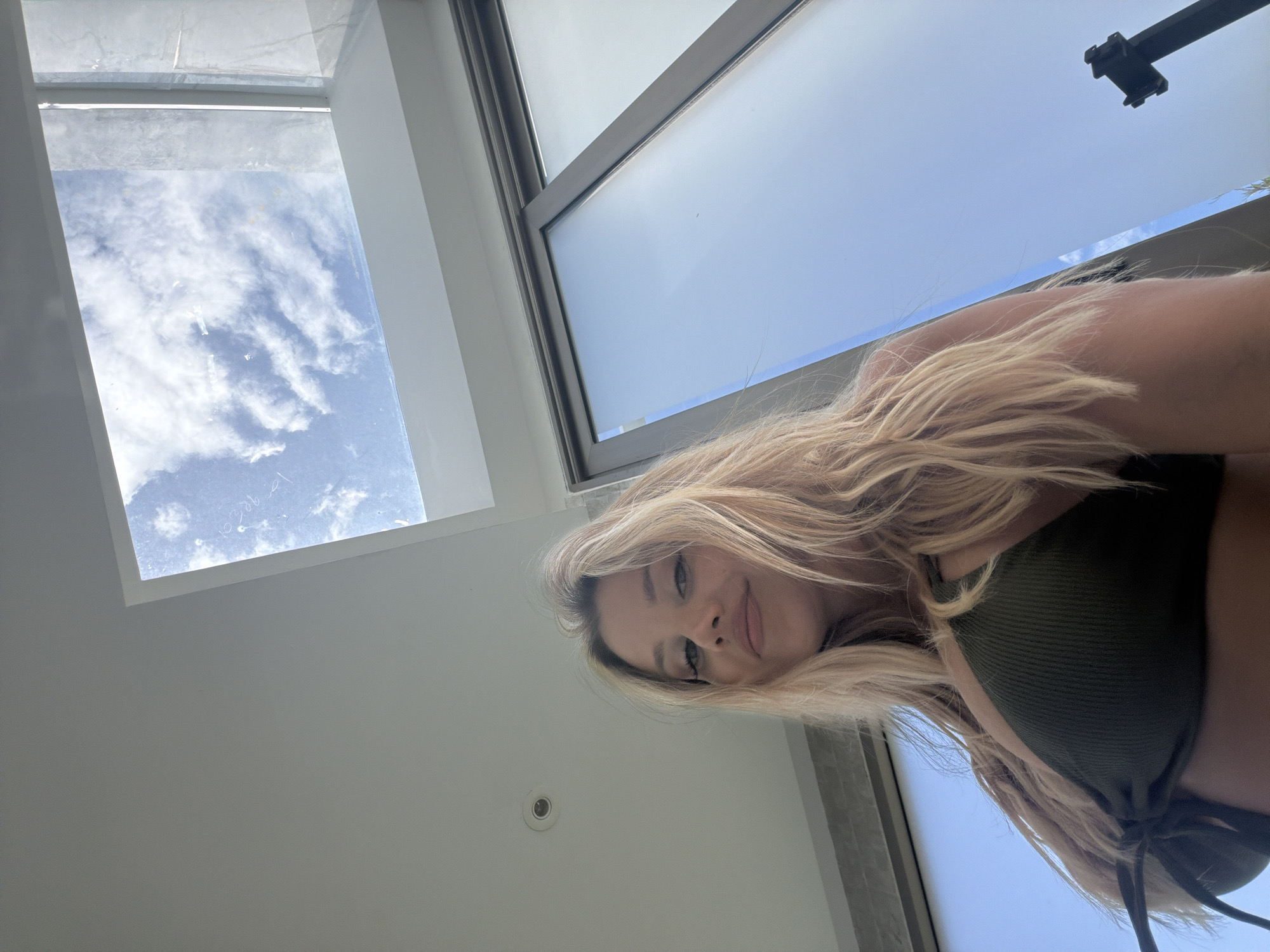In Thep Krasatti, Thailand, a young woman with blonde hair, wearing a dark green bikini top, is pictured from a low angle looking upwards. She is indoors, with a skylight above showing a blue sky with white clouds. To her right, a large window with frosted glass is visible, reflecting the blue sky. Part of a dark object, possibly a tripod, is seen in the lower right corner of the frame. The overall setting appears to be a modern interior with large windows and a skylight.