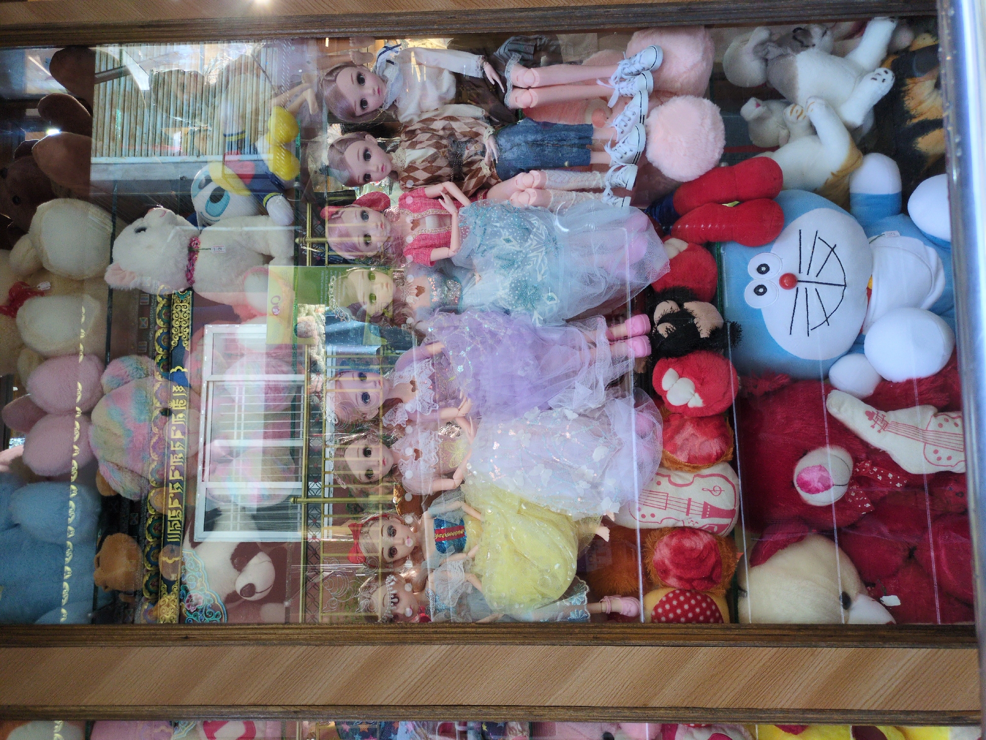 The image shows a display window filled with various stuffed toys, predominantly dolls and teddy bears. The dolls are arranged in rows, wearing elaborate dresses, with some standing and others seated. Behind the dolls, more stuffed toys are visible, including a white cat, a plush duck, and a large blue Doraemon character. The background appears to be a store interior with shelves and other merchandise. The display is framed by wooden elements, suggesting it is a shopfront. The overall impression is that of a toy store or a stall selling plush toys. While the image features many items, it is impossible to determine the specific location within Dharamshala, India, without additional context or identifying landmarks.