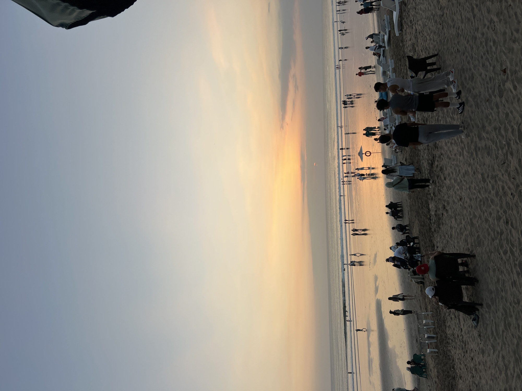 An aerial shot captures a wide view of Kuta Beach in Bali, Indonesia, during sunset. The sky displays a gradient of soft blues and oranges, with the sun peeking through a thin layer of clouds. The ocean, calm with gentle waves, stretches towards the horizon, reflecting the warm hues of the sky. A vast expanse of sand covers the foreground, populated by numerous people. Some are seen walking along the shoreline, while others are scattered across the beach, some sitting in chairs and others standing in groups. Beach chairs and umbrellas are visible, along with a few objects resembling coolers. The overall scene depicts a lively and serene beach atmosphere during the golden hour.